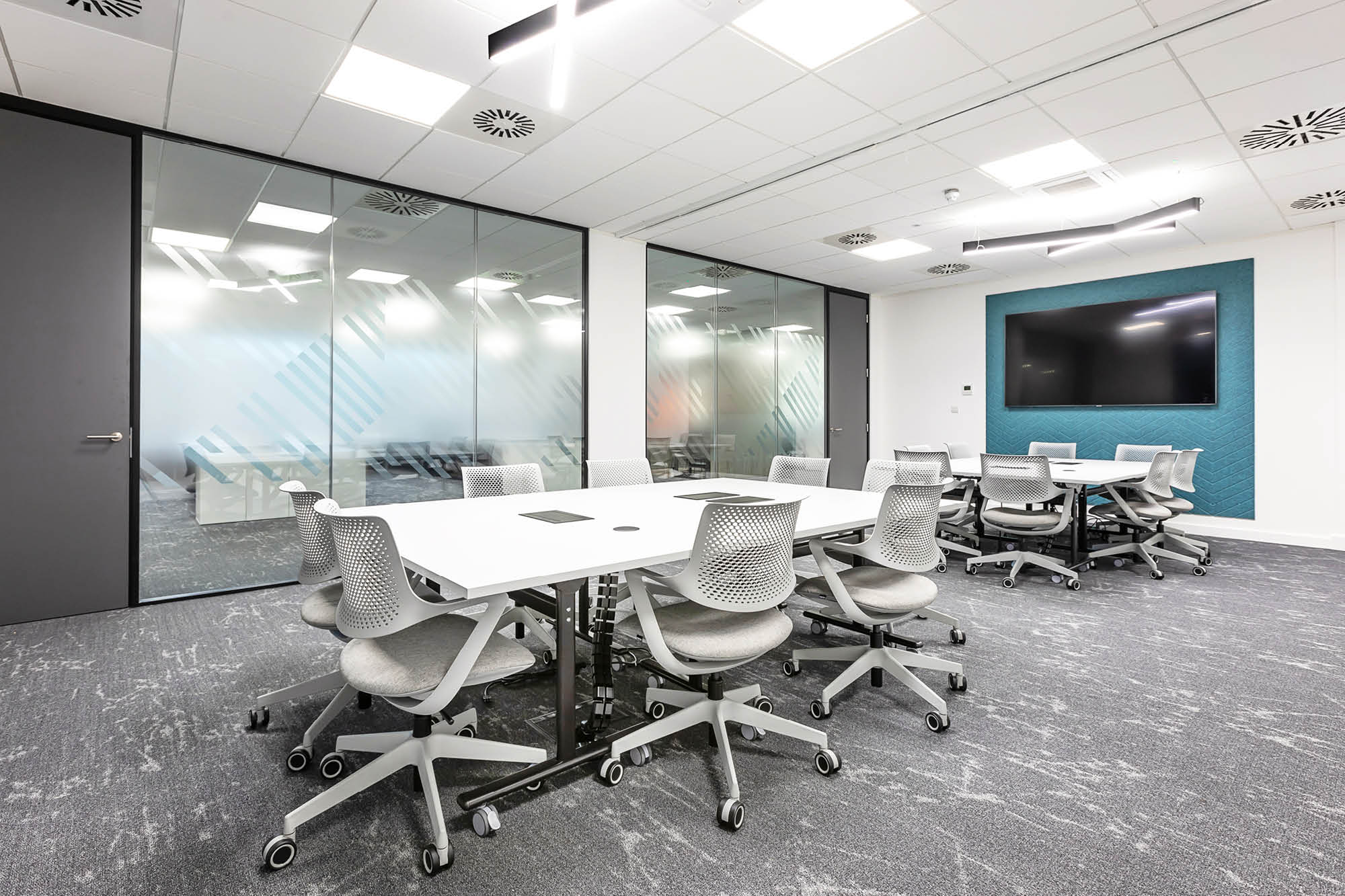 A modern conference room with white tables arranged in a U-shape. The room has grey rolling chairs, a large wall-mounted screen.