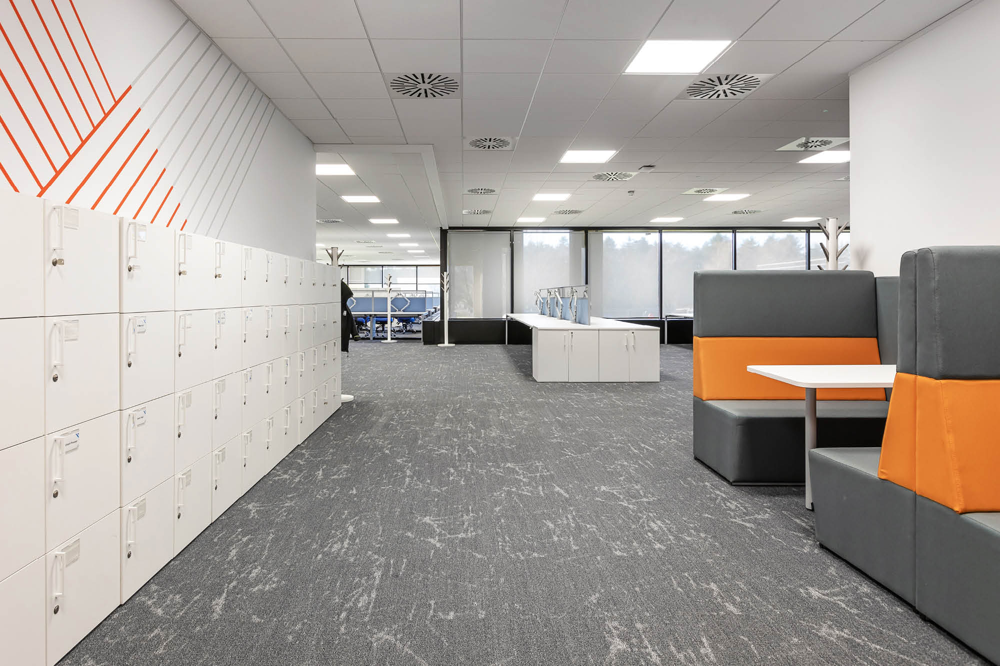 Modern office space with white lockers on the left and grey-and-orange booths on the right. The floor is carpeted in grey.