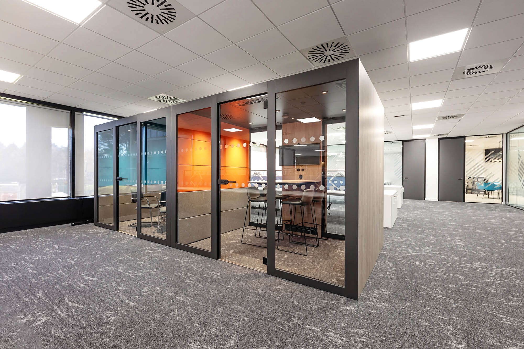 Modern office space featuring a glass-enclosed meeting room with orange and blue accent walls. Inside, theres a table with chairs.