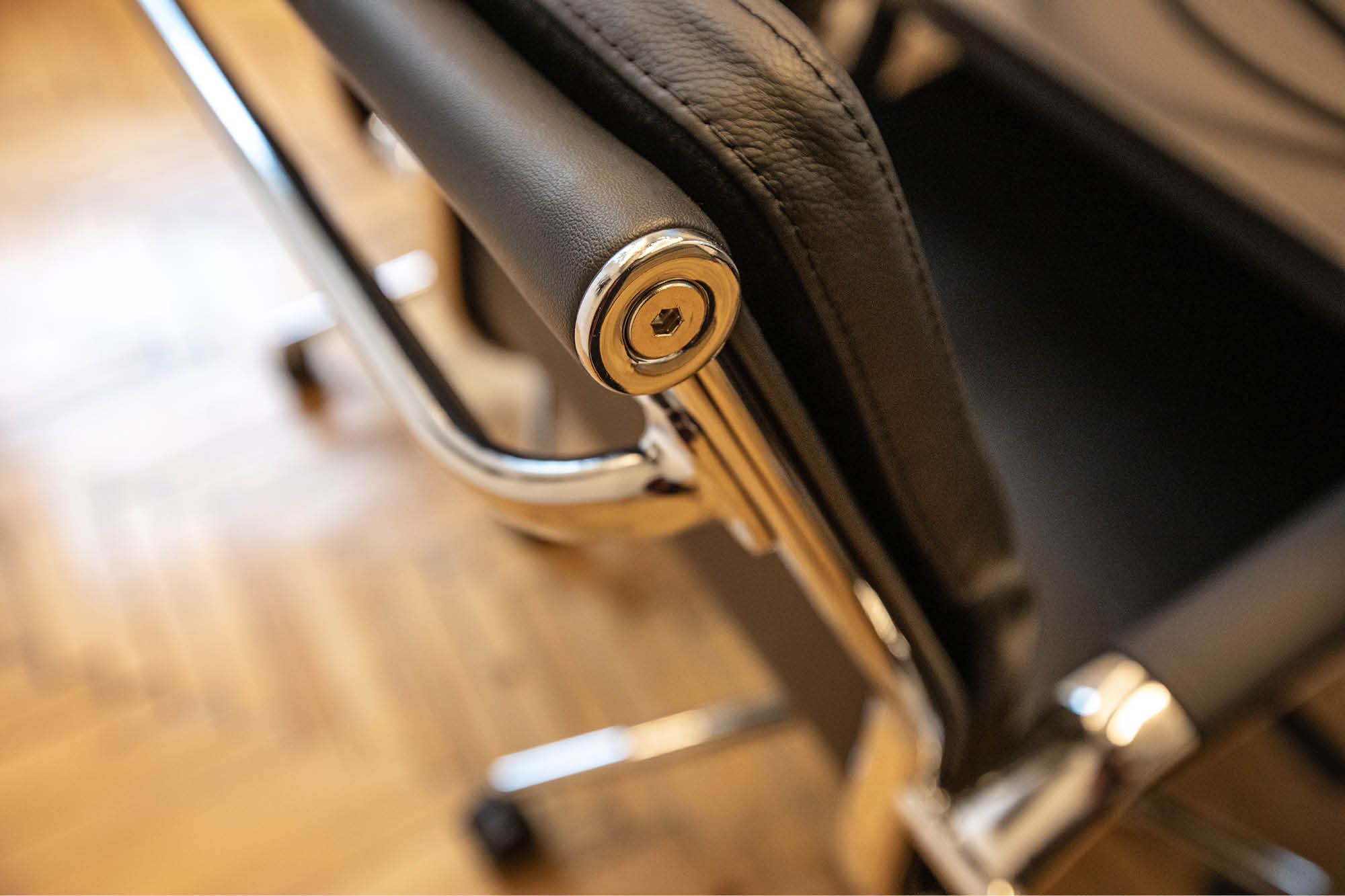 Close-up of a black leather office chair with shiny metal armrests. The metal frame reflects light and the chair is positioned on a wooden floor.