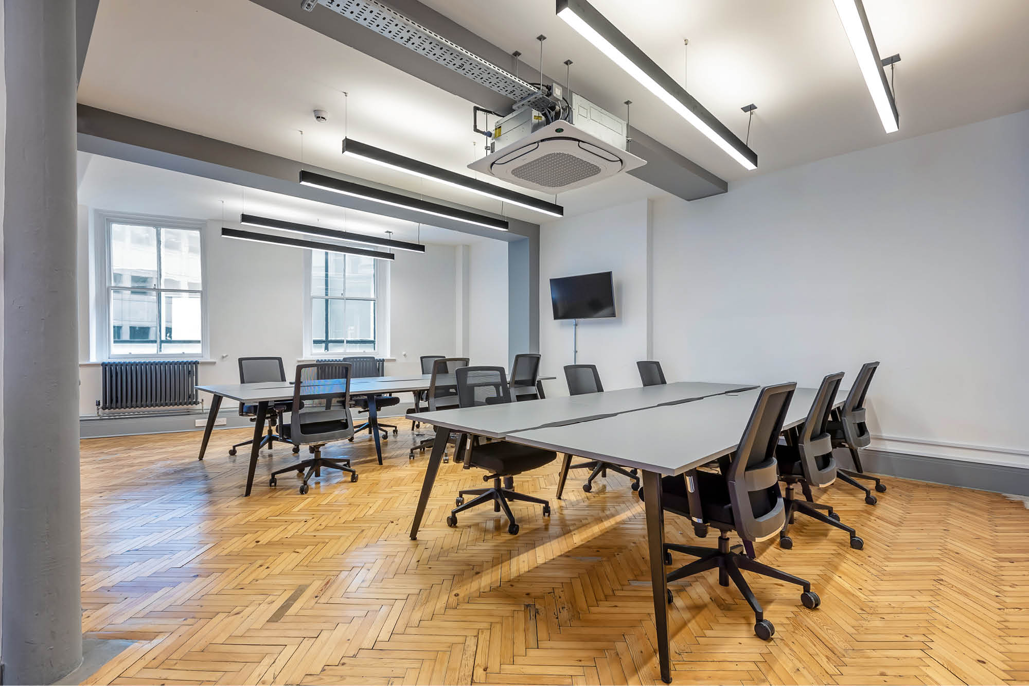 A modern office meeting room with wooden herringbone flooring. It features two large tables arranged in a U-shape.