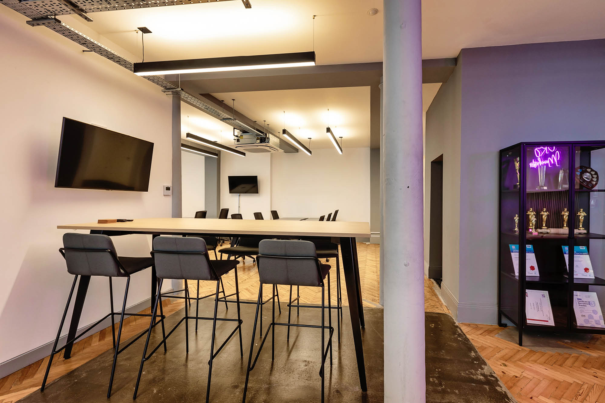Modern office space with a high conference table surrounded by bar stools, two wall-mounted flat-screen TVs.
