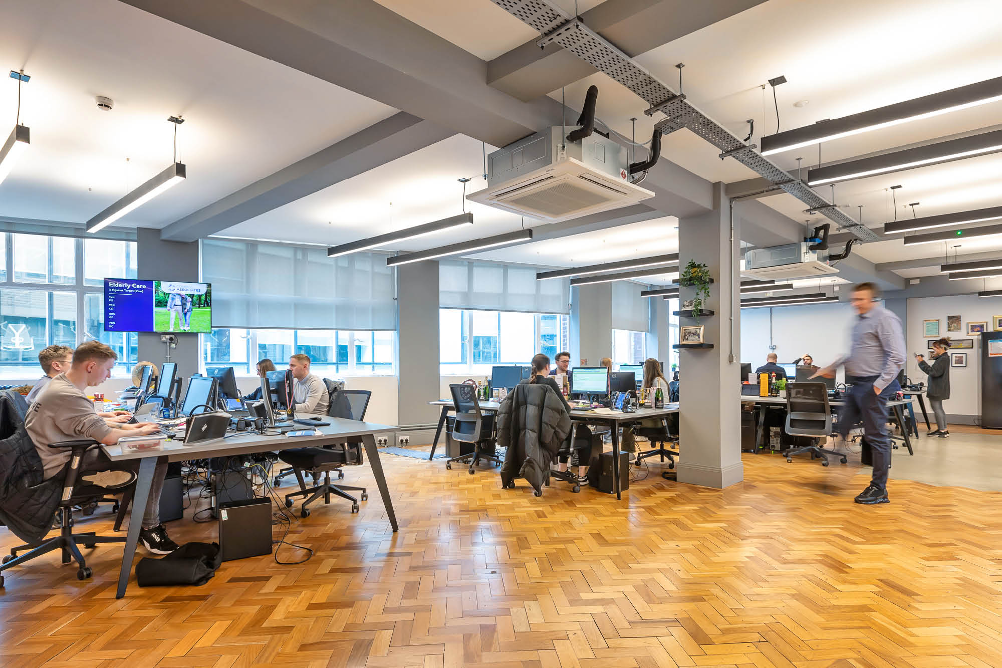 A modern office space with several people working at desks. The room features a herringbone wood floor, large windows with blinds.