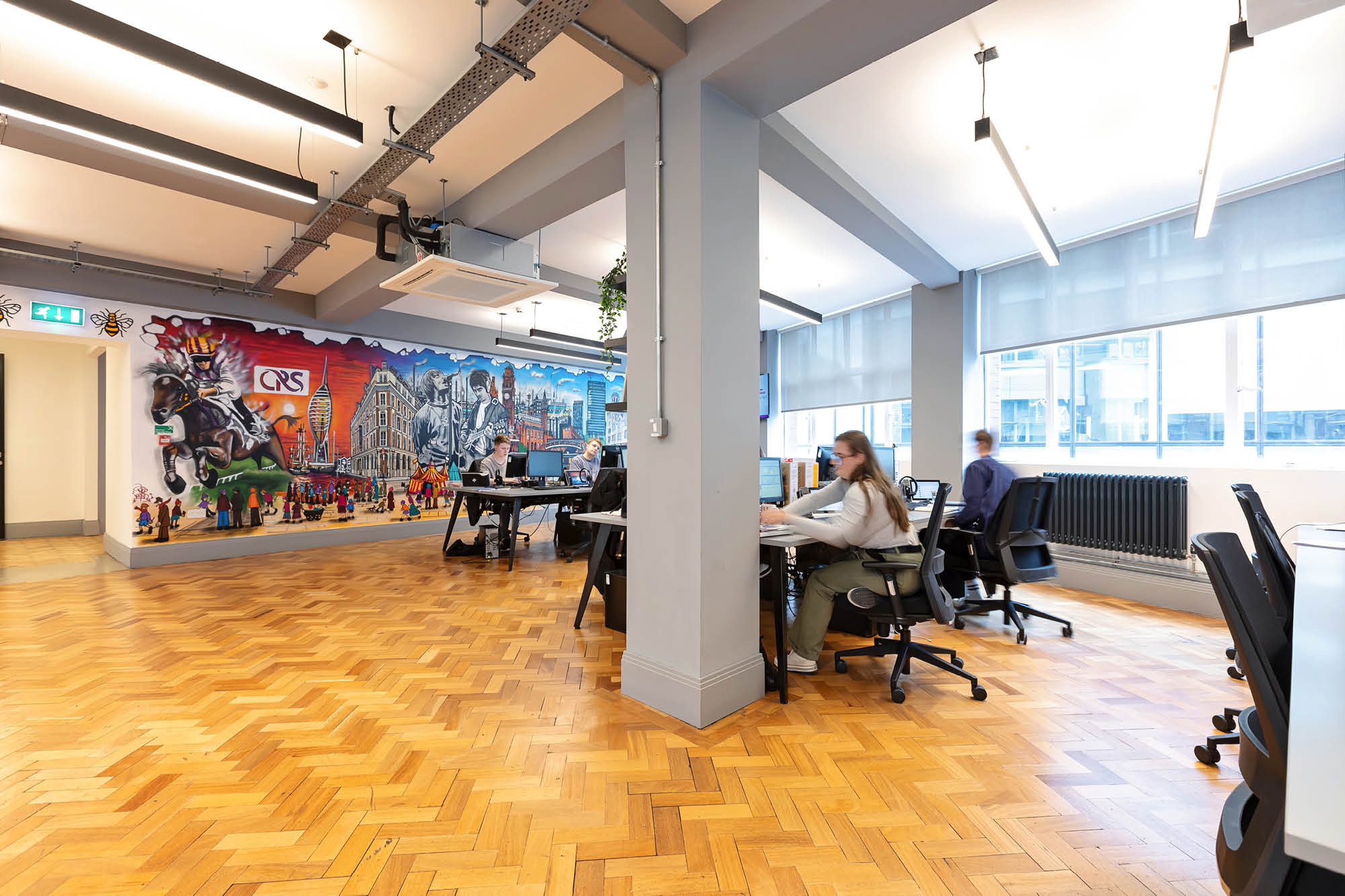 Office space with wooden herringbone flooring, featuring desks with people working on computers.