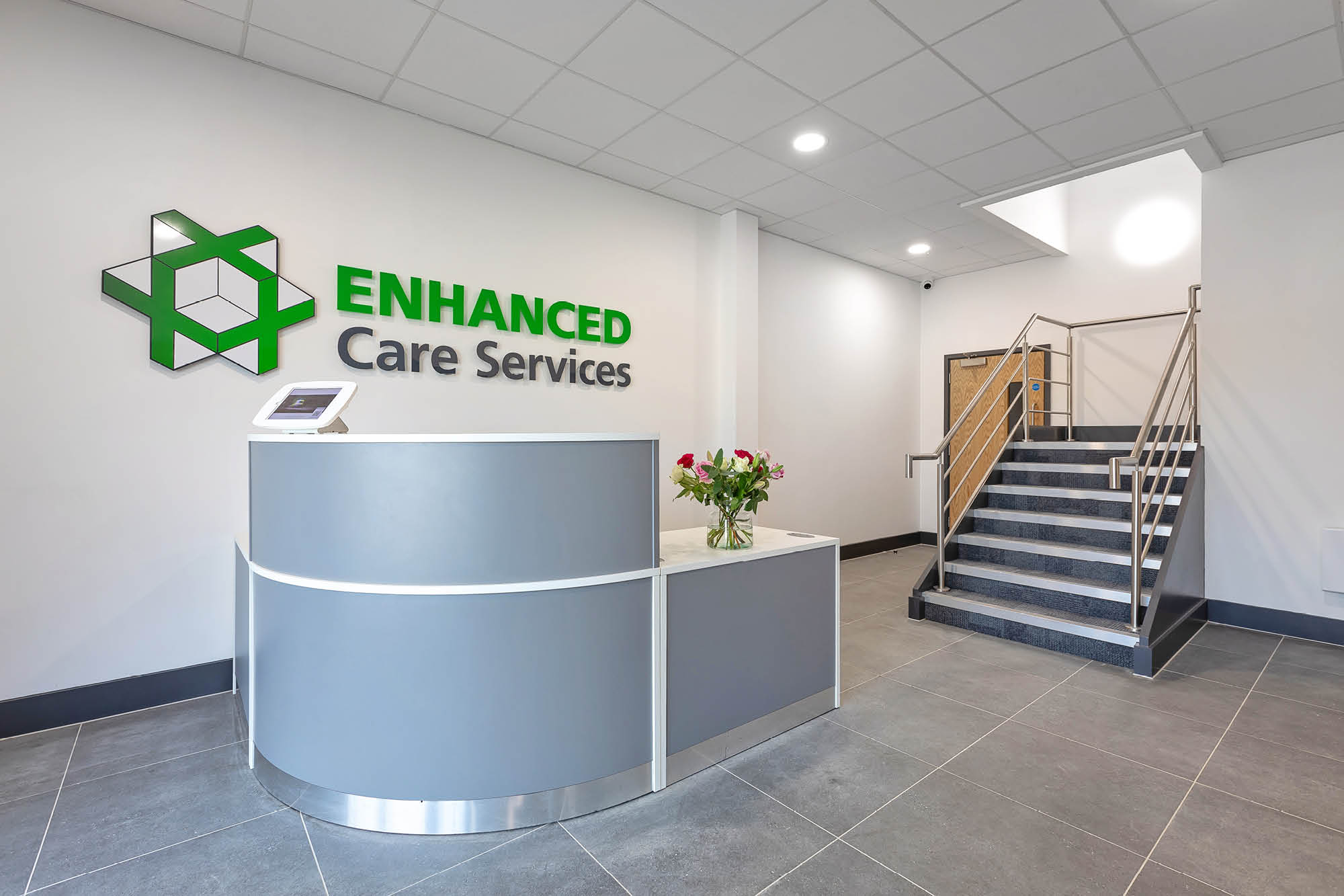 The image shows a modern office reception area with a curved grey desk displaying flowers and a tablet. A sign on the wall reads ENHANCED Care Services.