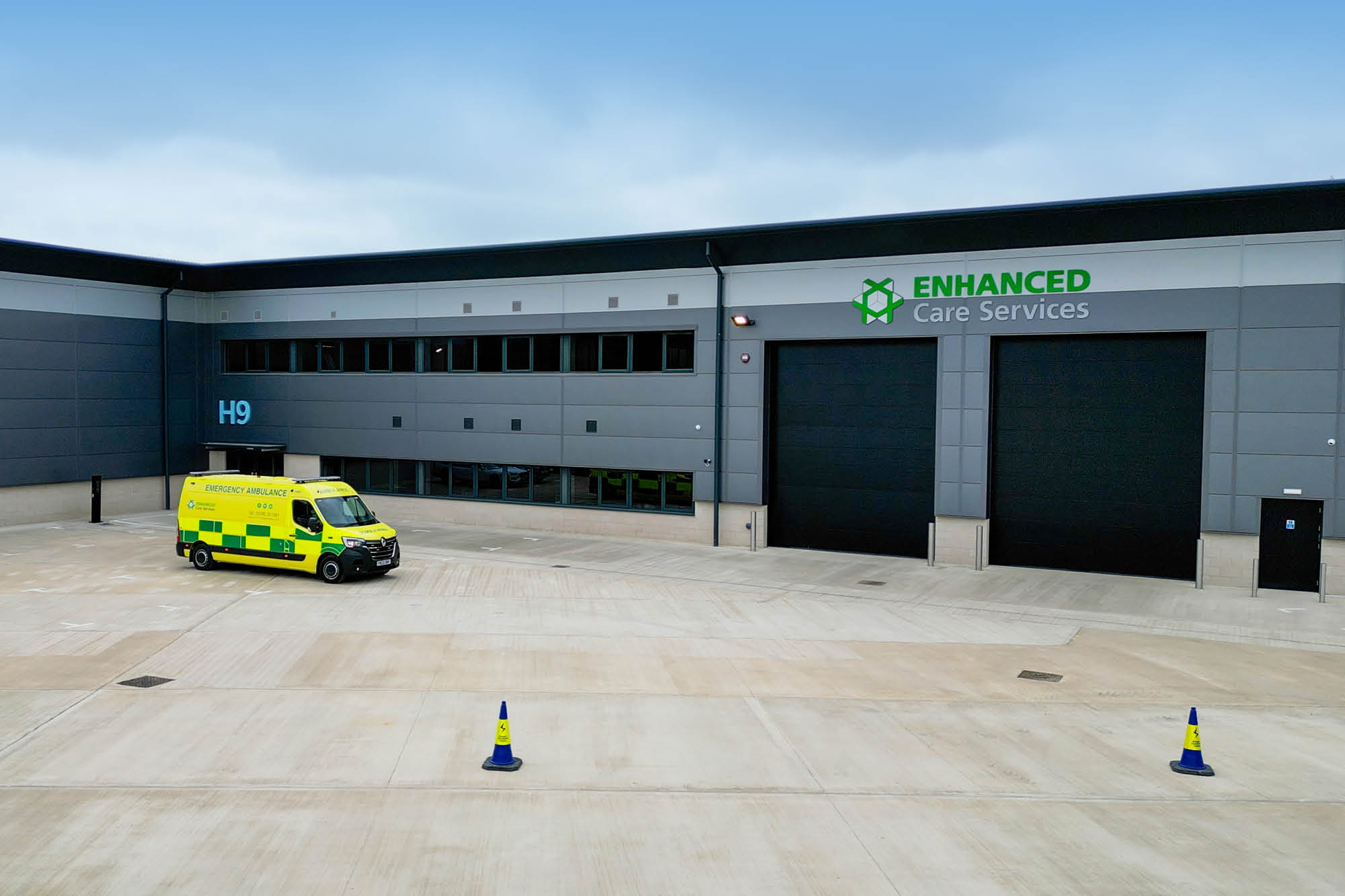 A bright yellow ambulance is parked outside a large, modern warehouse building labeled ENHANCED Care Services and marked H9.