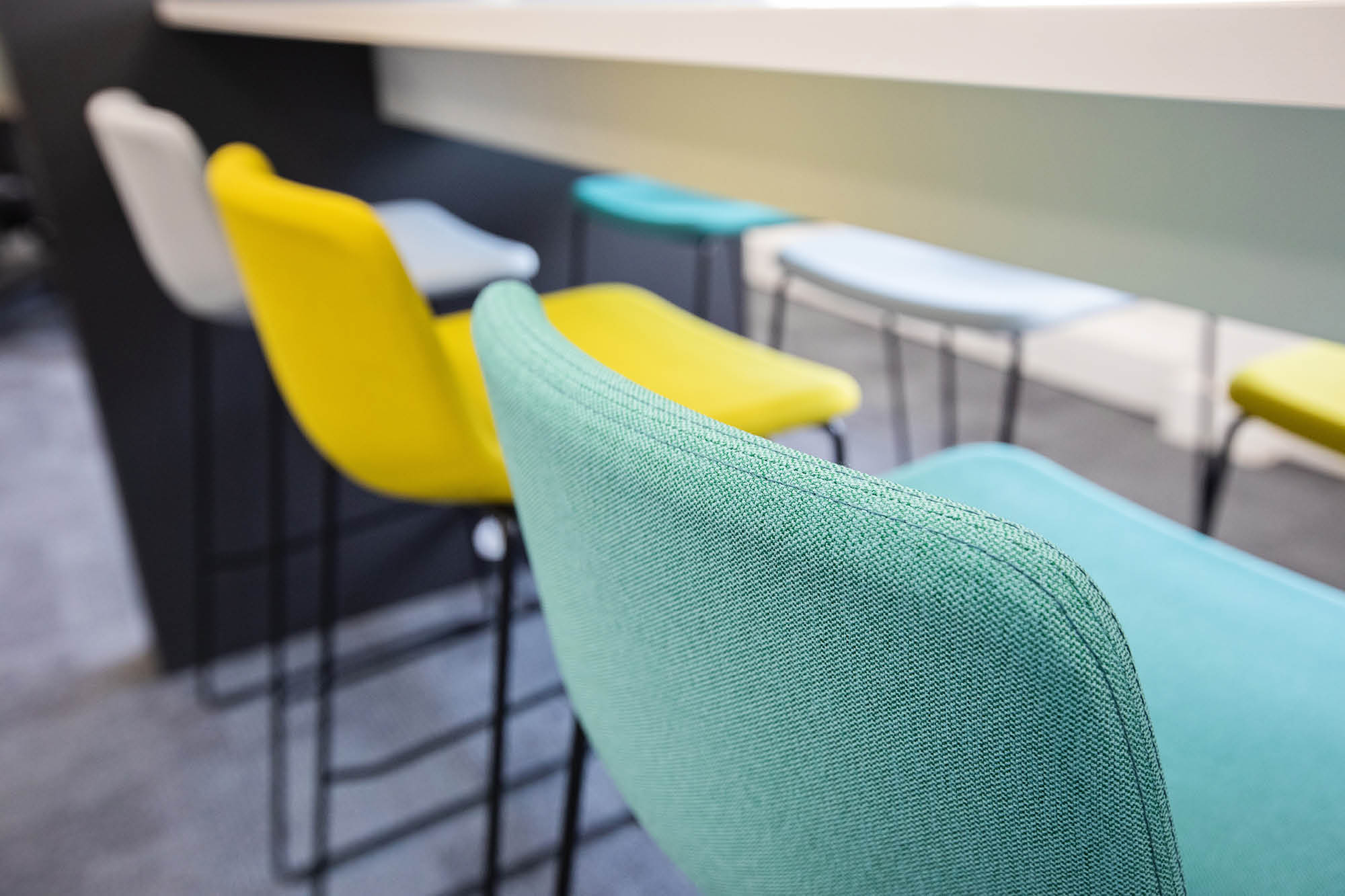 Colorful bar stools with fabric upholstery placed along a counter. The stools are in shades of turquoise, yellow, and blue, with black metal legs.