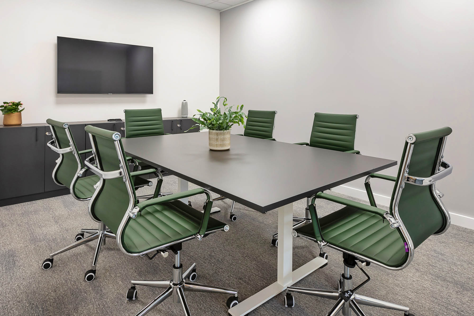 A modern conference room with a black rectangular table surrounded by six green swivel chairs. A flat-screen TV is mounted on the wall.