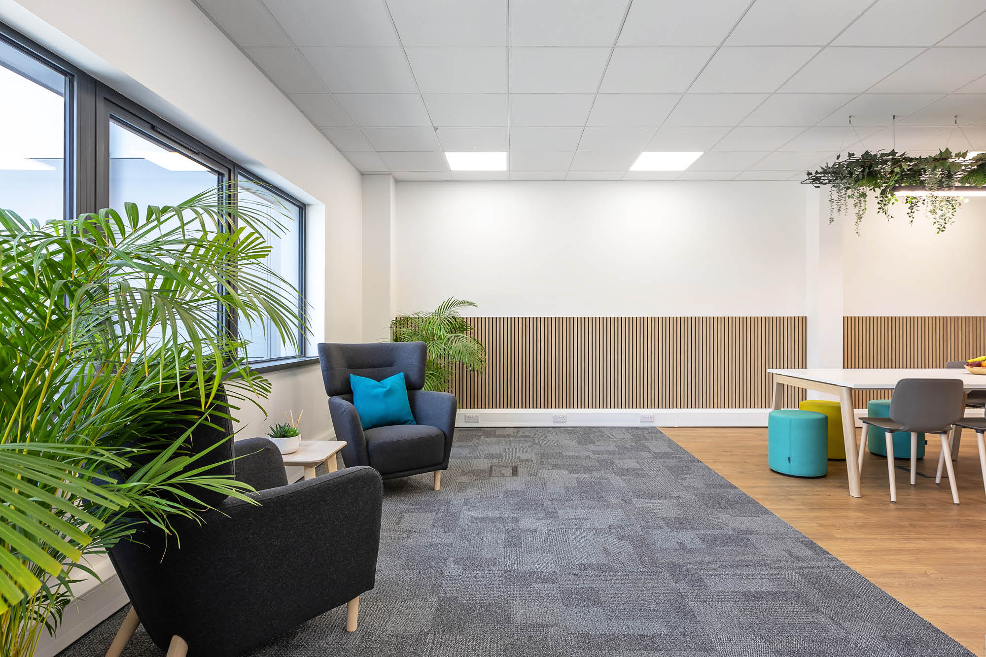 Modern office waiting area with gray armchairs, a small round table, and a blue cushion. A large window provides natural light.