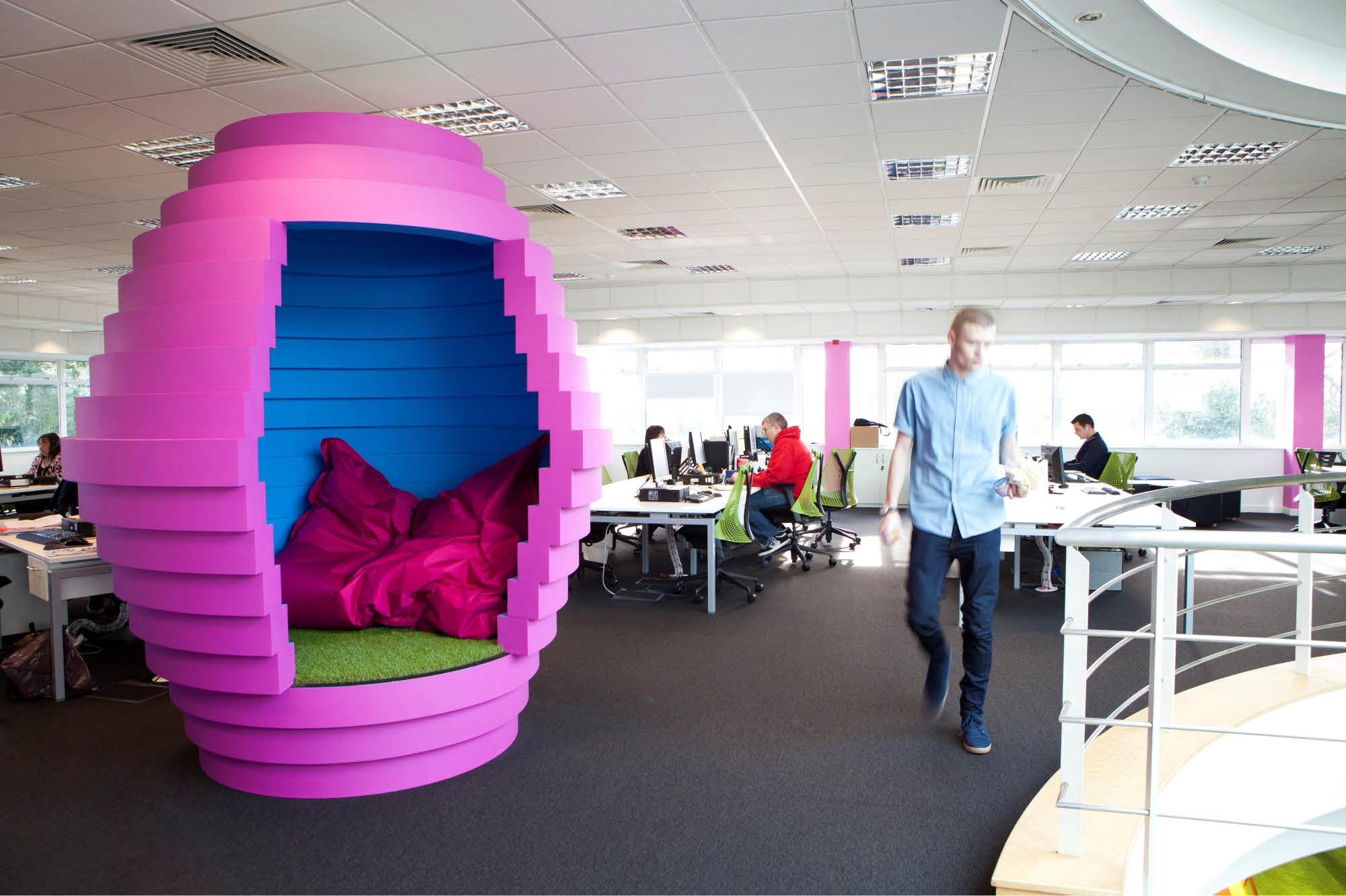 A modern office space featuring a unique, egg-shaped, pink and blue pod with a person walking by.