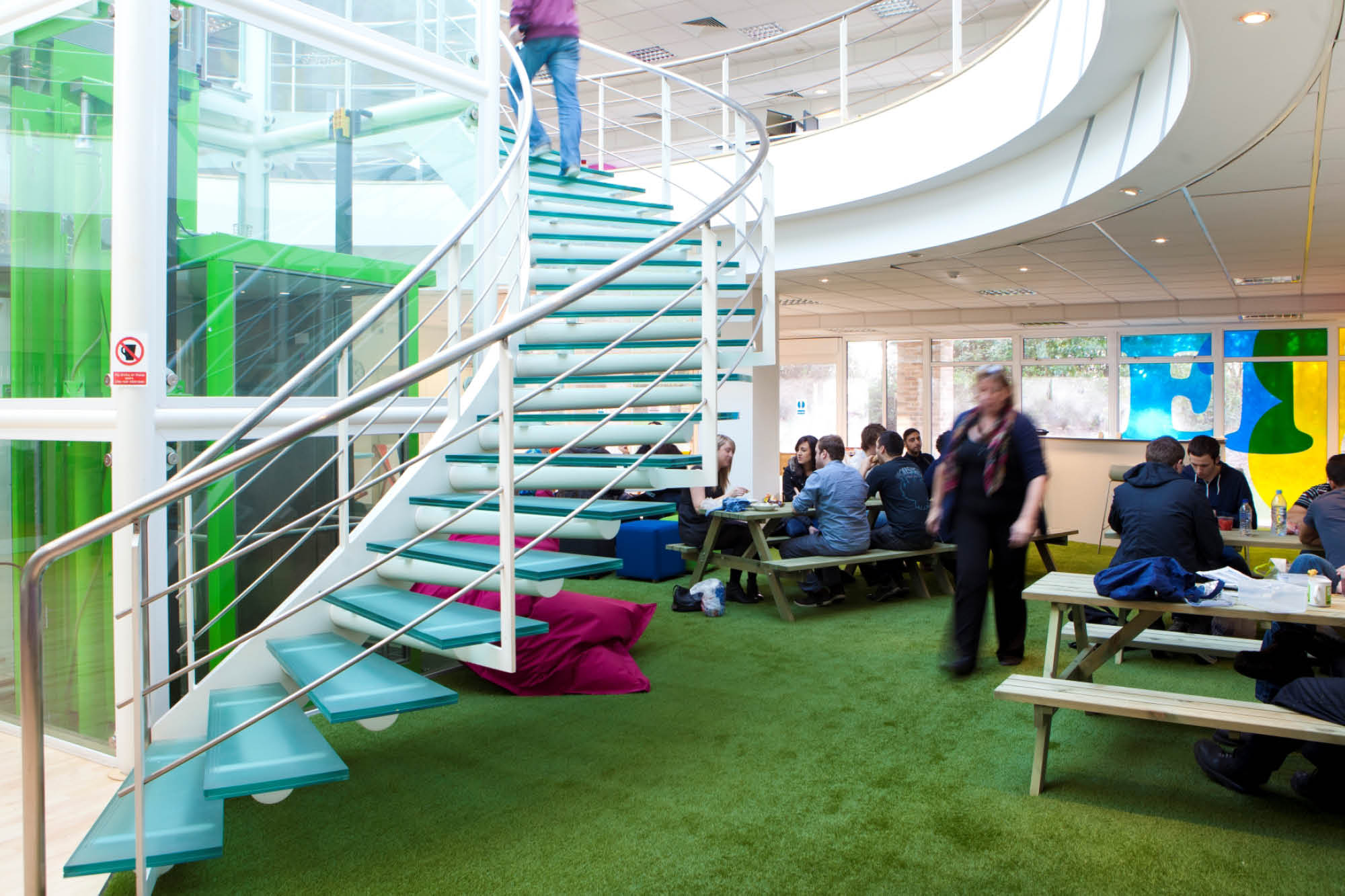 A modern office interior with green carpeting features a spiral staircase on the left. Several people are sitting at picnic tables.