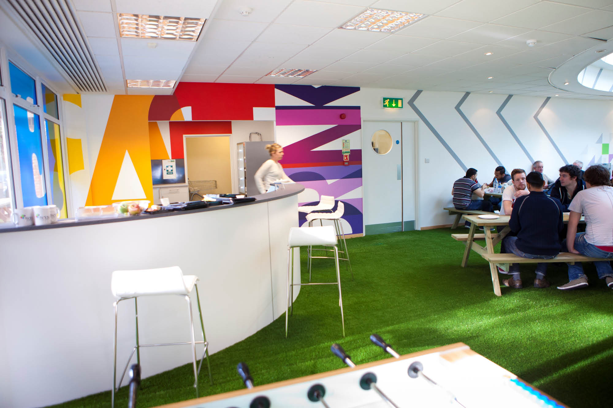 A modern office break room features colourful wall art and green carpet resembling grass.