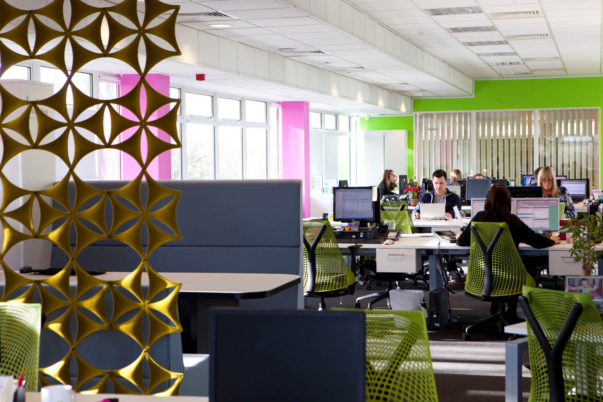 Open office space with bright green chairs, white desks, and pink and green accents. Several people work at computers.