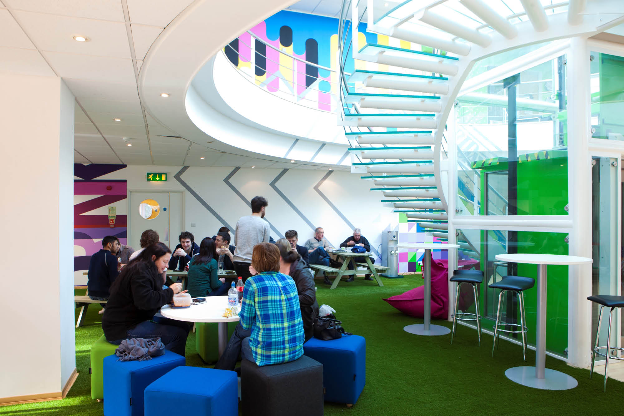 A modern office lounge with people sitting and conversing at tables. The space features artificial grass flooring.