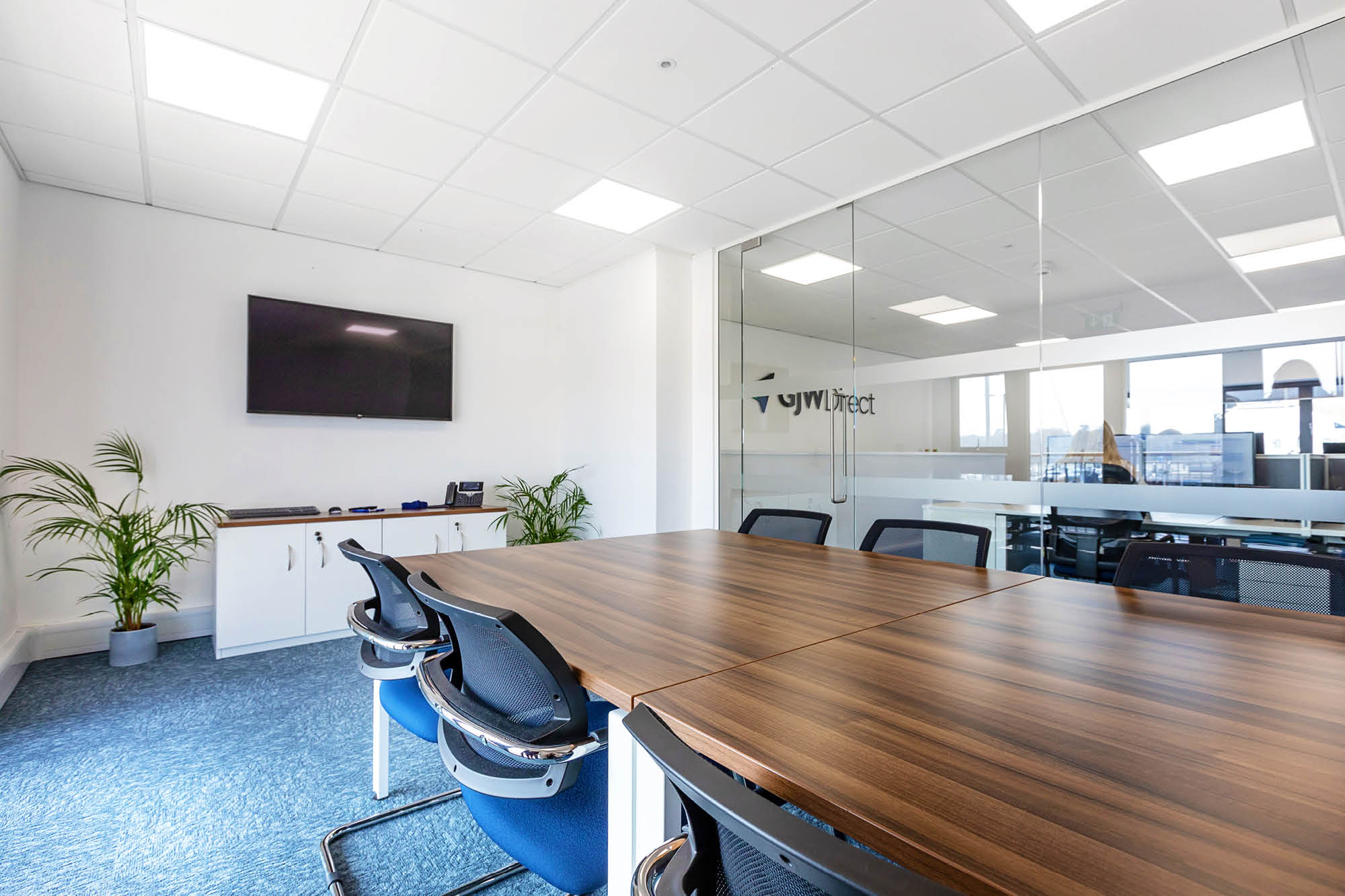 A modern conference room with a large wooden table surrounded by blue office chairs. The room has a wall-mounted TV, potted plants.