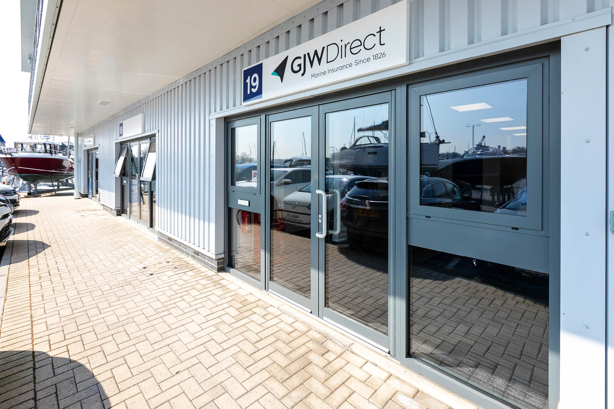 Exterior of GJW Direct office with large glass doors and signage. The building is light gray with a brick walkway.