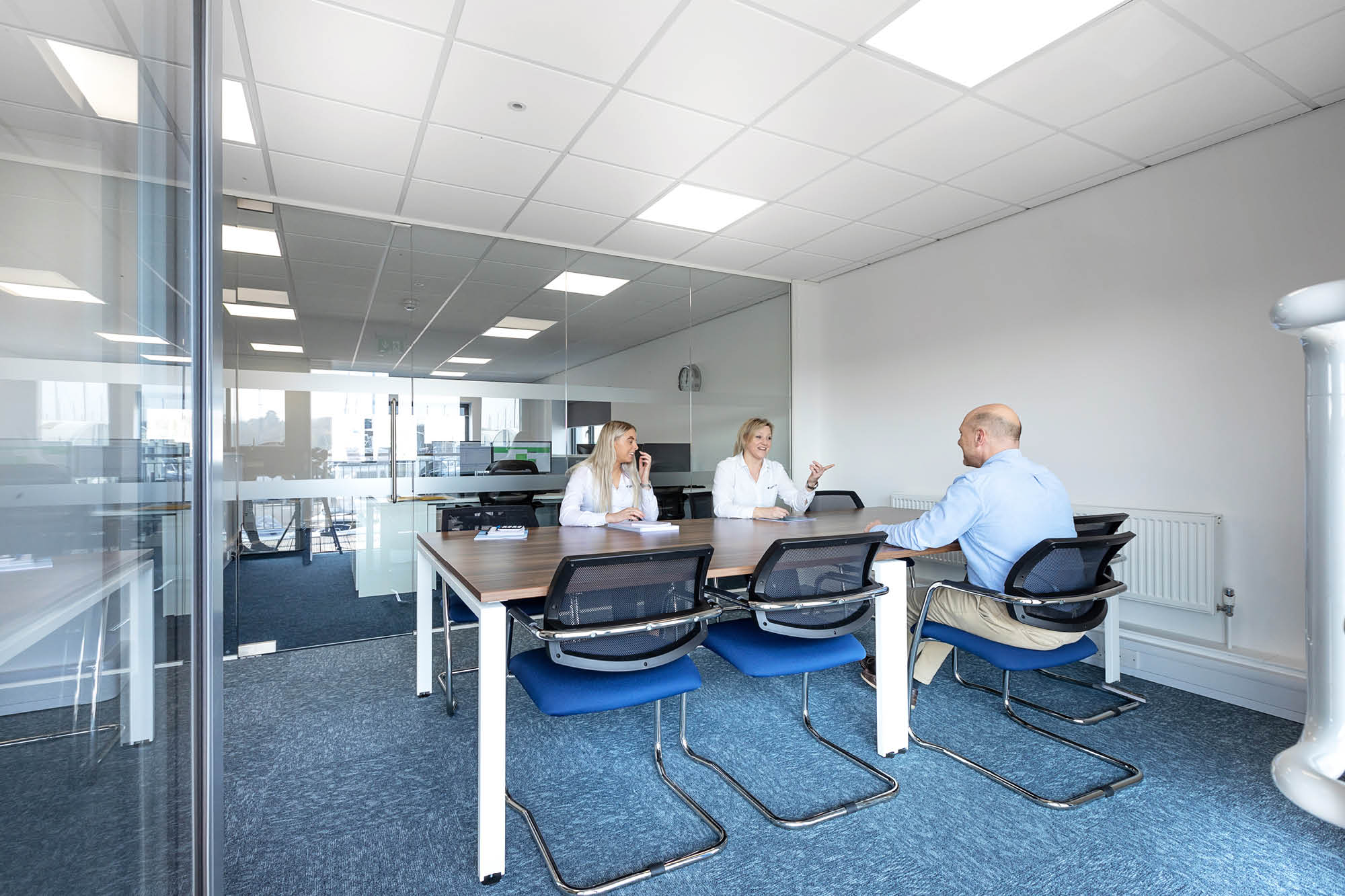 Three people are in a glass-walled conference room with blue carpet and a rectangular table. Two women and a man are engaged in conversation.