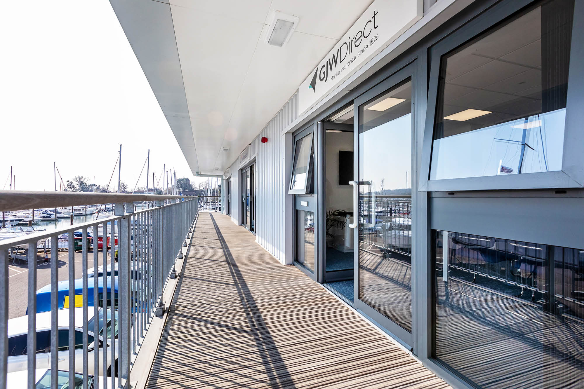 Exterior view of an office with large windows and a glass door on a wooden deck, overlooking a marina filled with boats.
