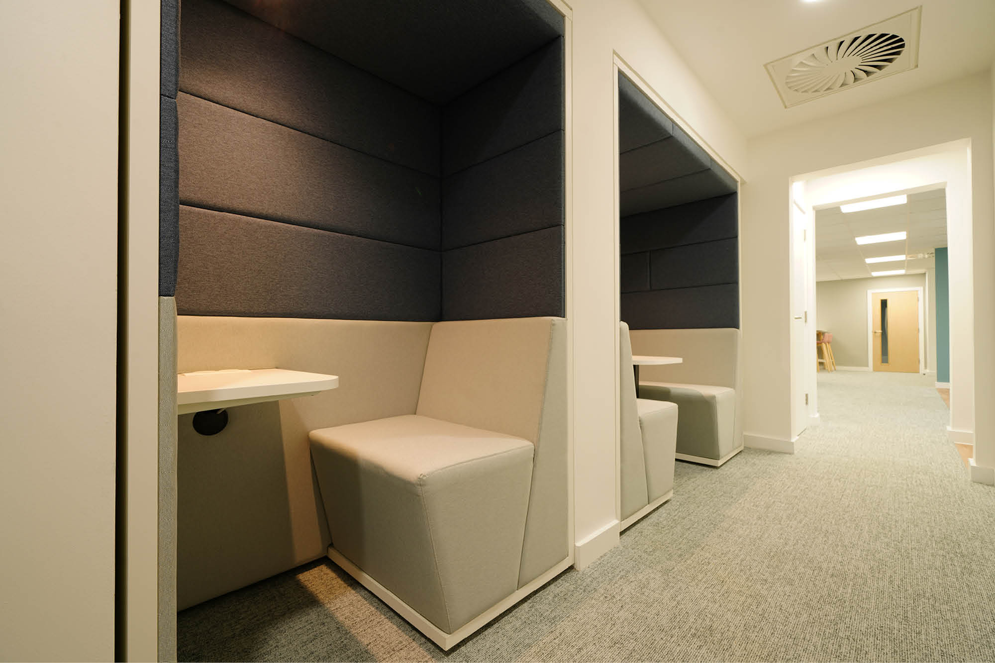 A hallway with individual seating booths featuring cushioned walls and small tables. The area is illuminated by overhead lighting.
