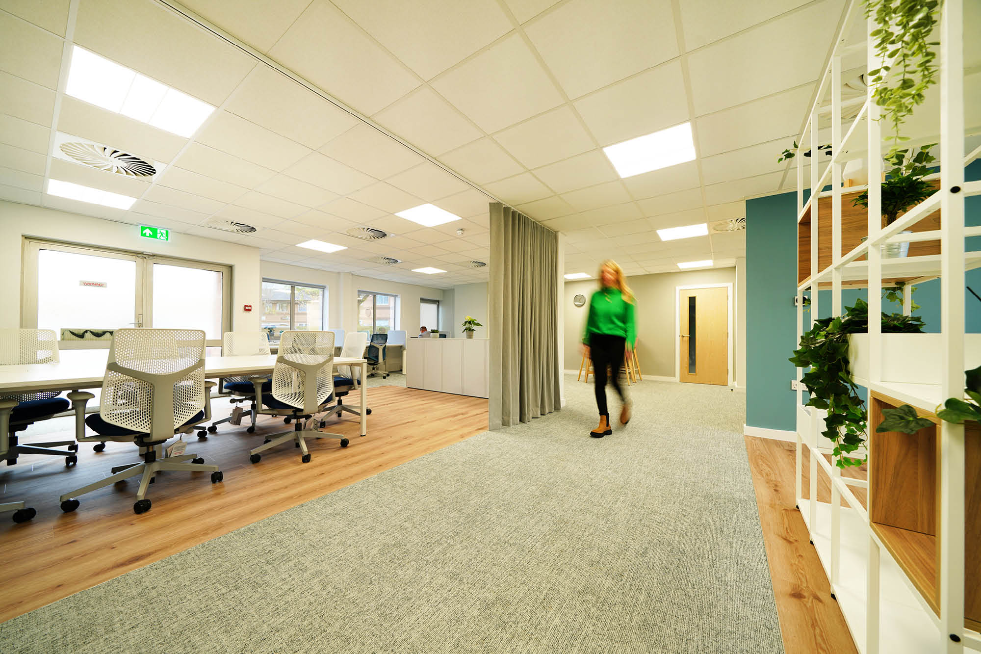 A modern office space with a person walking past. The room features long desks with ergonomic chairs.