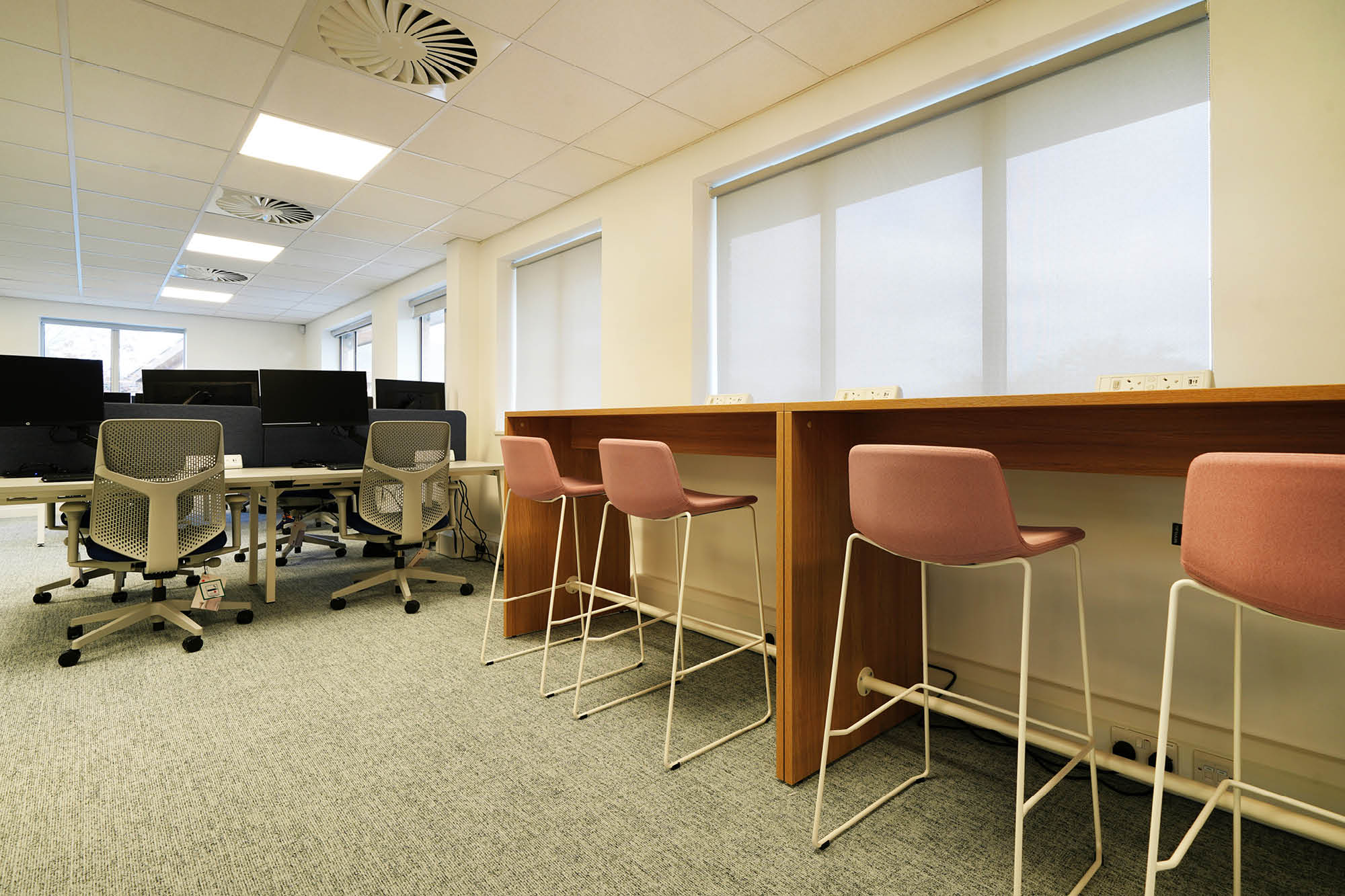 Modern office space with long wooden table and four pink barstools on the right. On the left, several grey office.