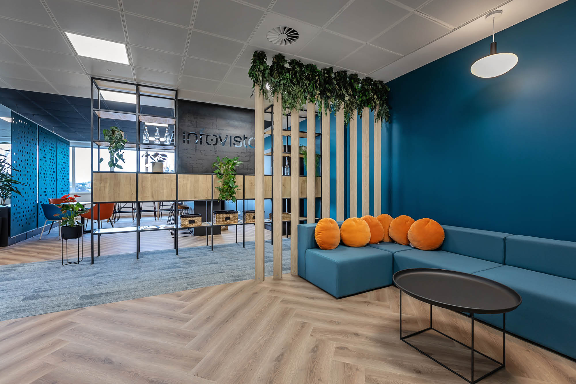 Modern office lounge with teal walls, wooden flooring, and a blue sectional sofa featuring orange cushions.
