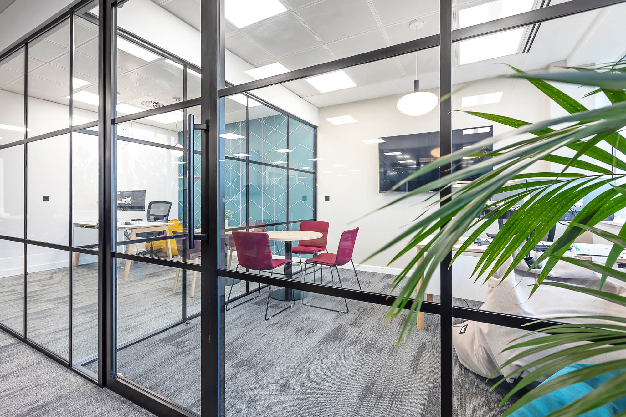 Modern office space with glass walls showing a small meeting area with a round table and three pink chairs.