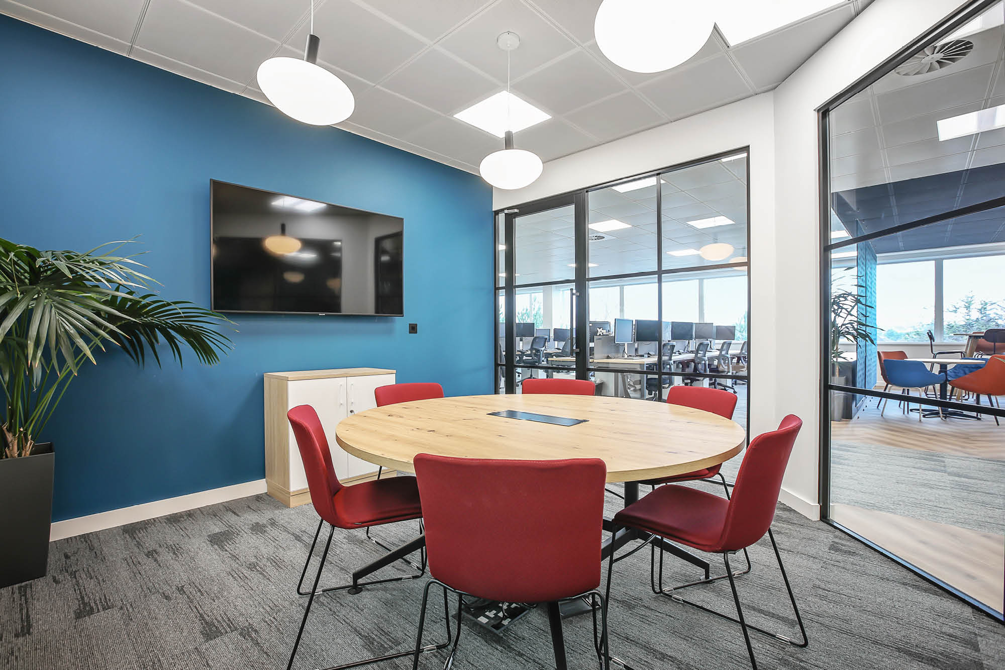 A modern meeting room with a round wooden table, red chairs, and a large flat-screen TV mounted on a blue wall.
