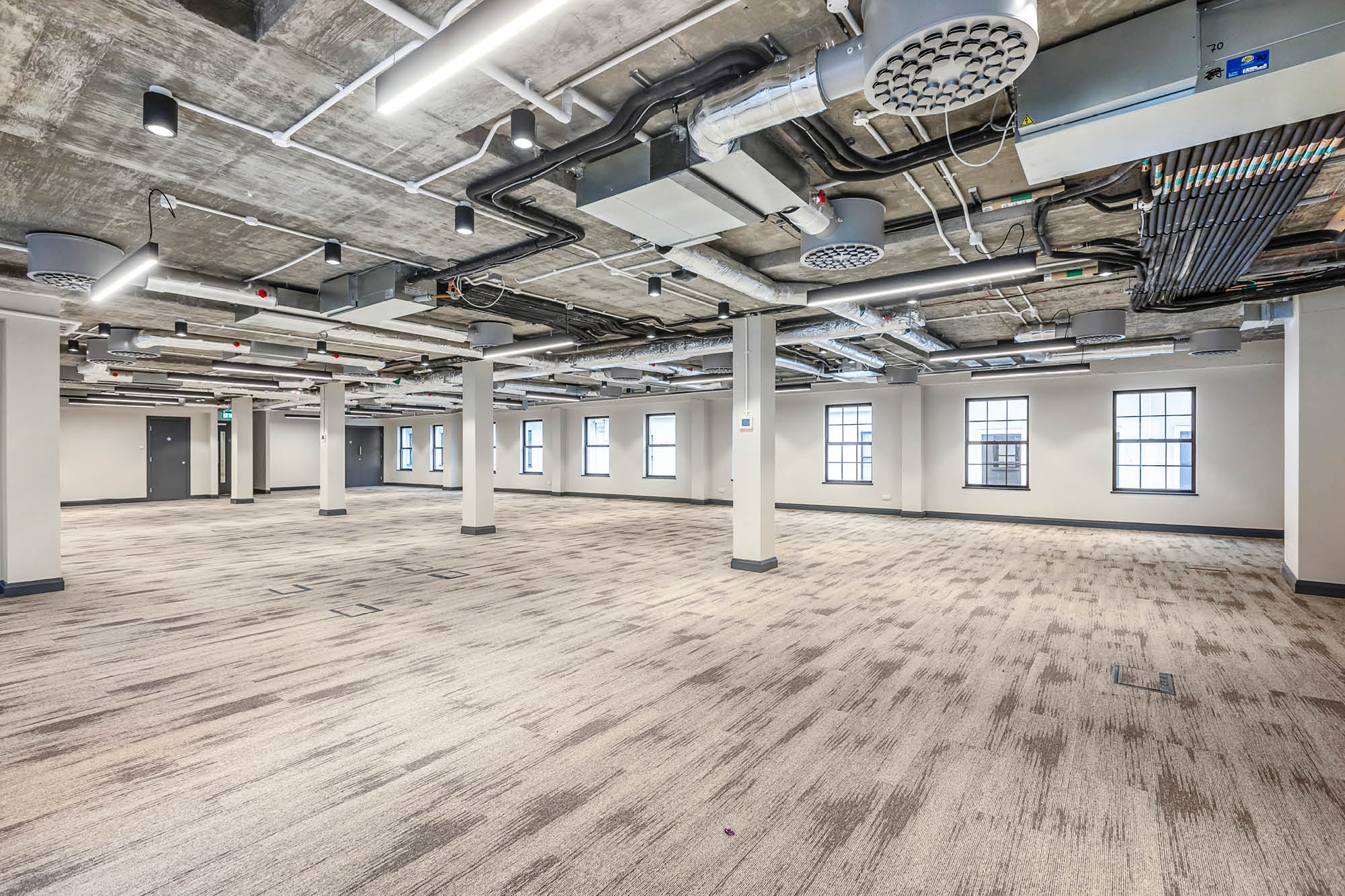 A spacious, industrial-style open office area with exposed ceilings, ductwork, fluorescent lighting.