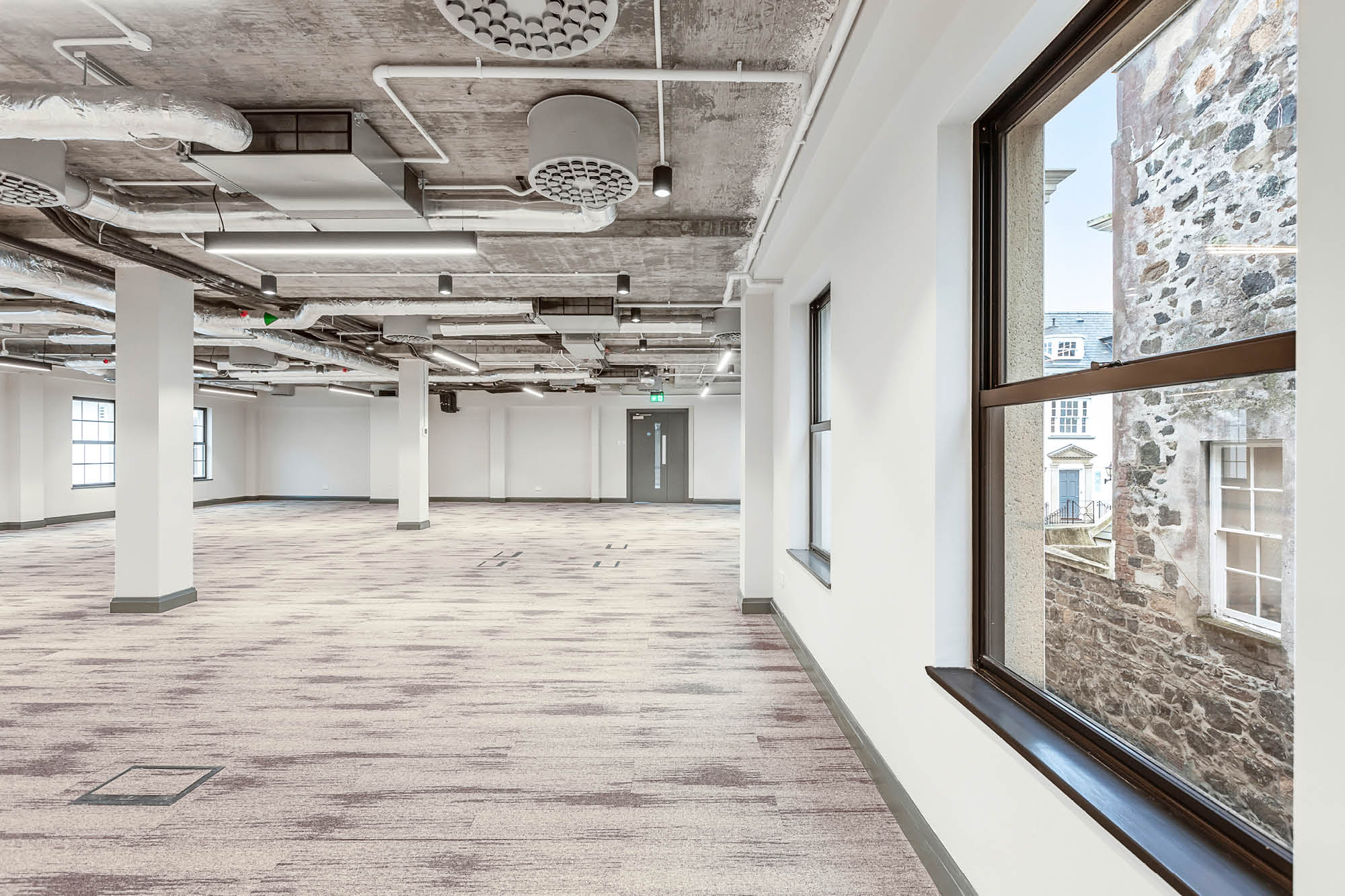 Spacious, empty office interior with large windows, white walls, exposed ceiling pipes and fixtures.