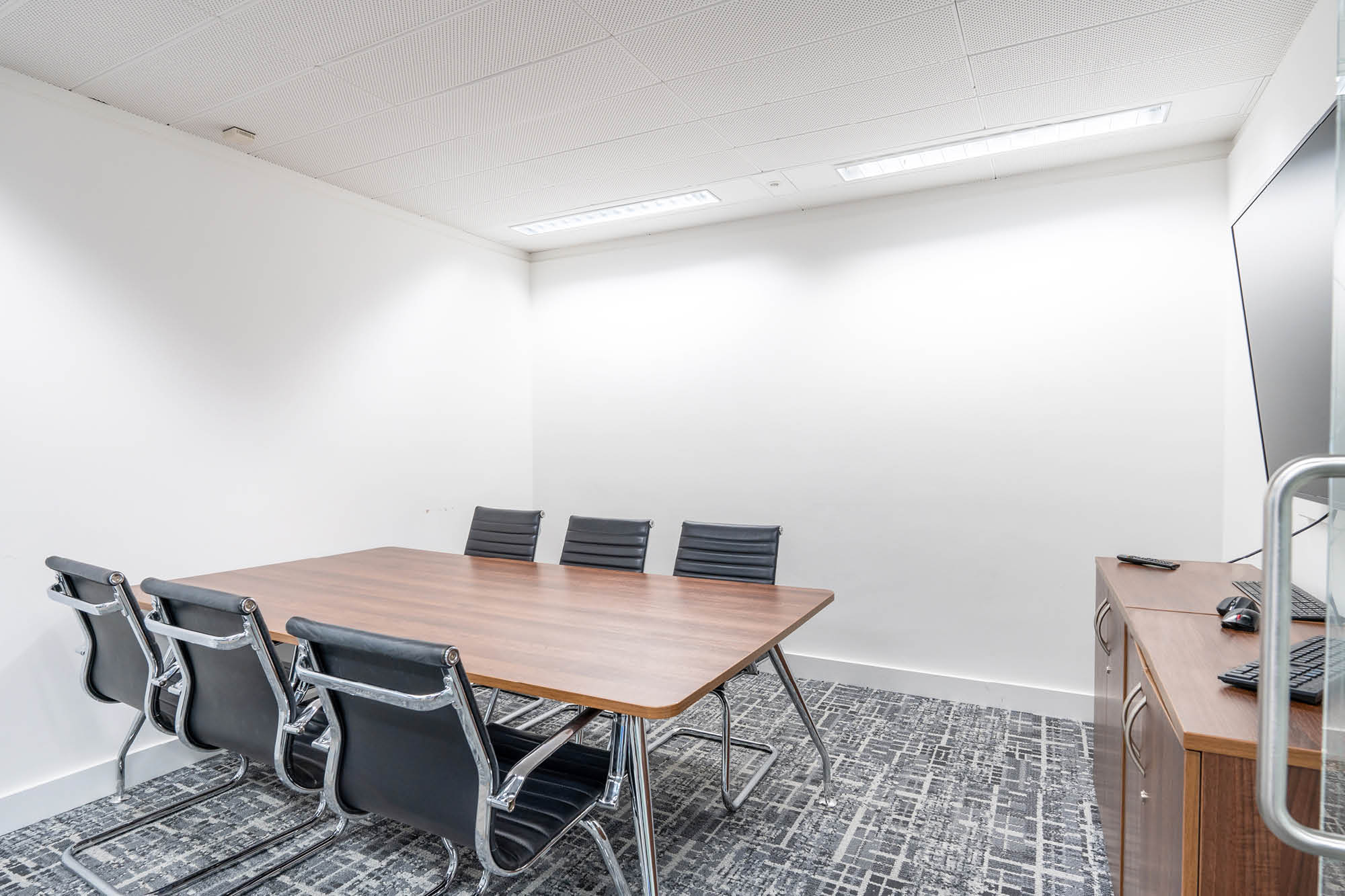 A modern conference room with a wooden table and six black chairs. The room has white walls and a patterned gray carpet.