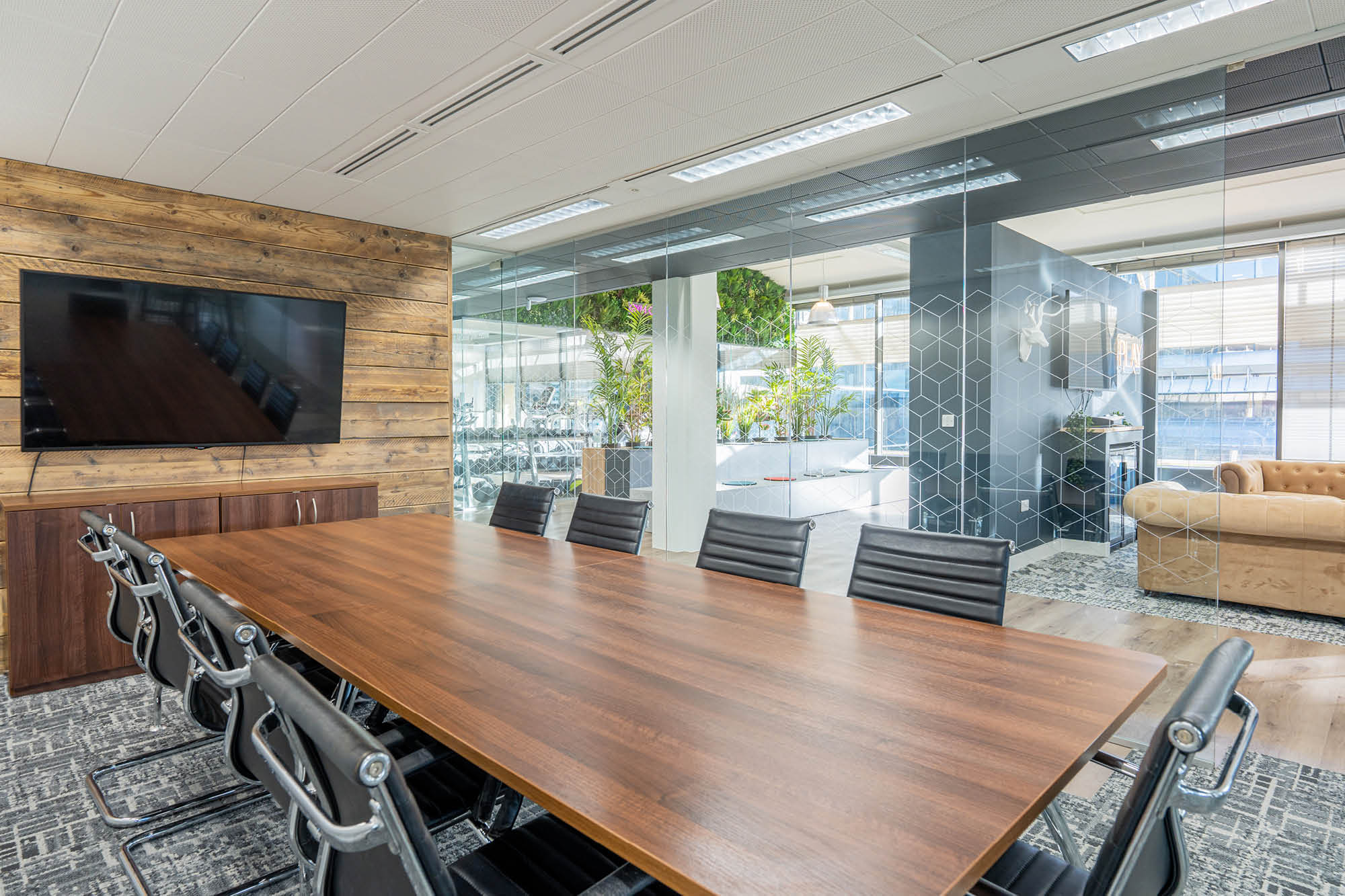 Modern conference room with a long wooden table and black leather chairs. A large TV is mounted on a wooden accent wall.