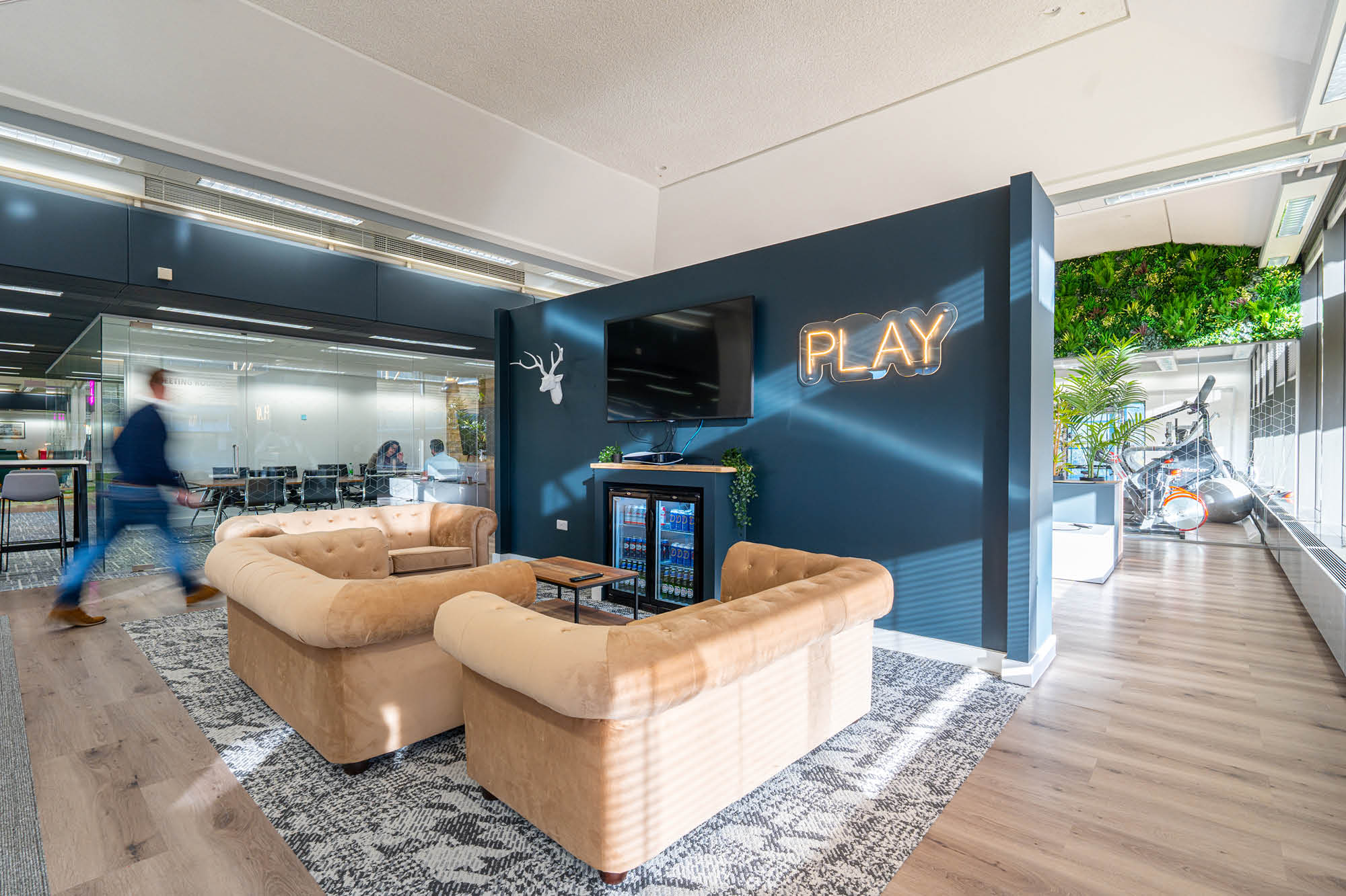 Modern office lounge area with two beige sofas and a small table on a patterned rug. A dark blue wall features a mounted TV.