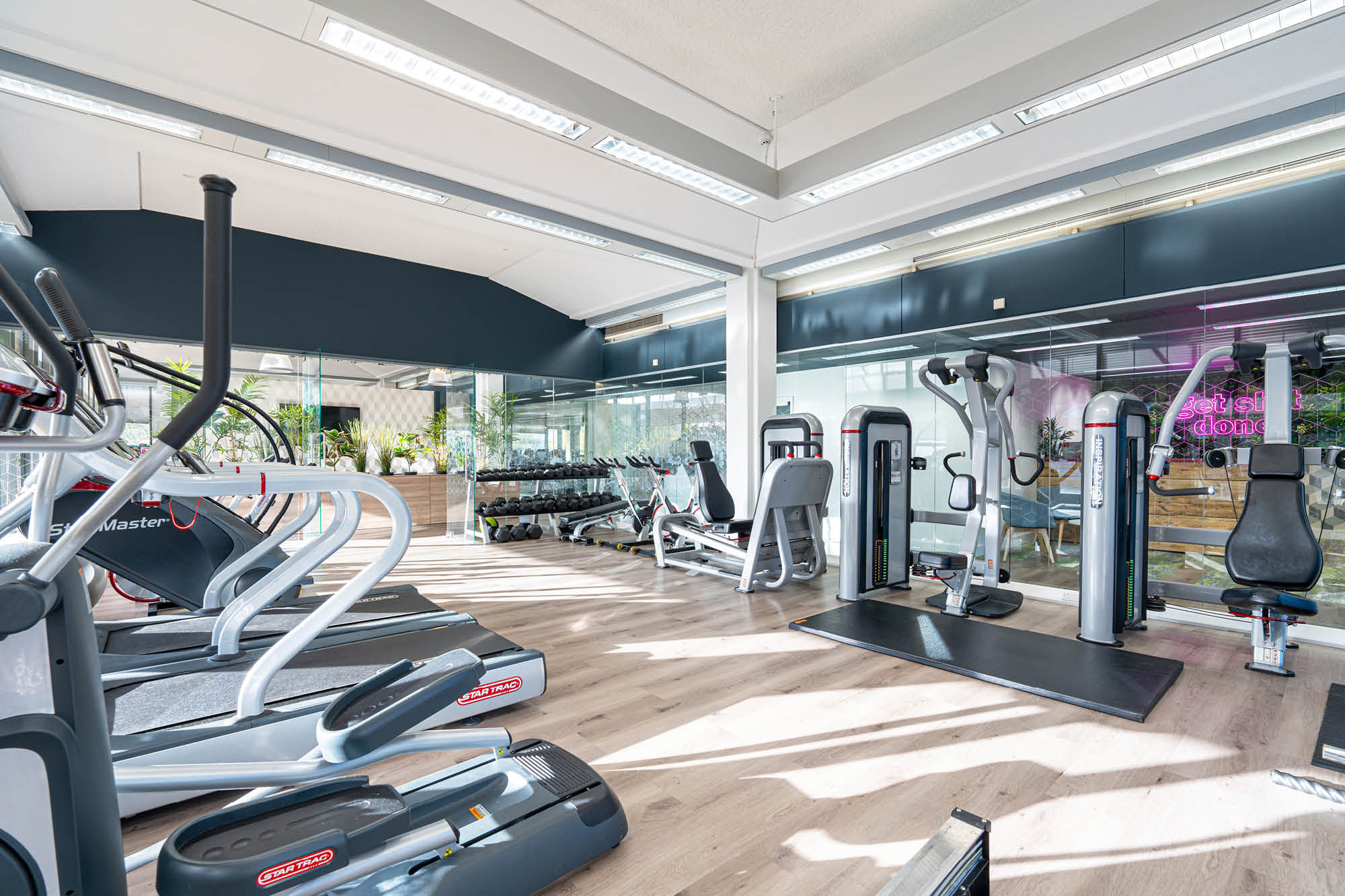 Modern gym with exercise equipment, including treadmills, elliptical machines, and weight machines. Bright space with large windows.