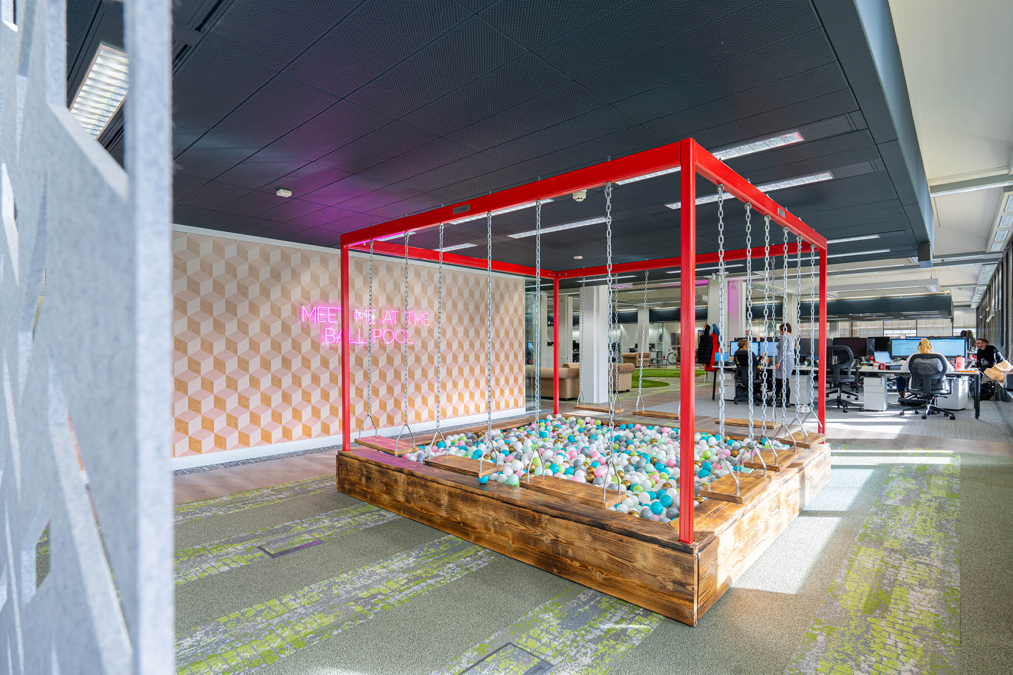 Open office space with a ball pit featuring swings suspended over it. A neon sign on a geometric-patterned wall reads MEET ME AT THE PLAYPEN.