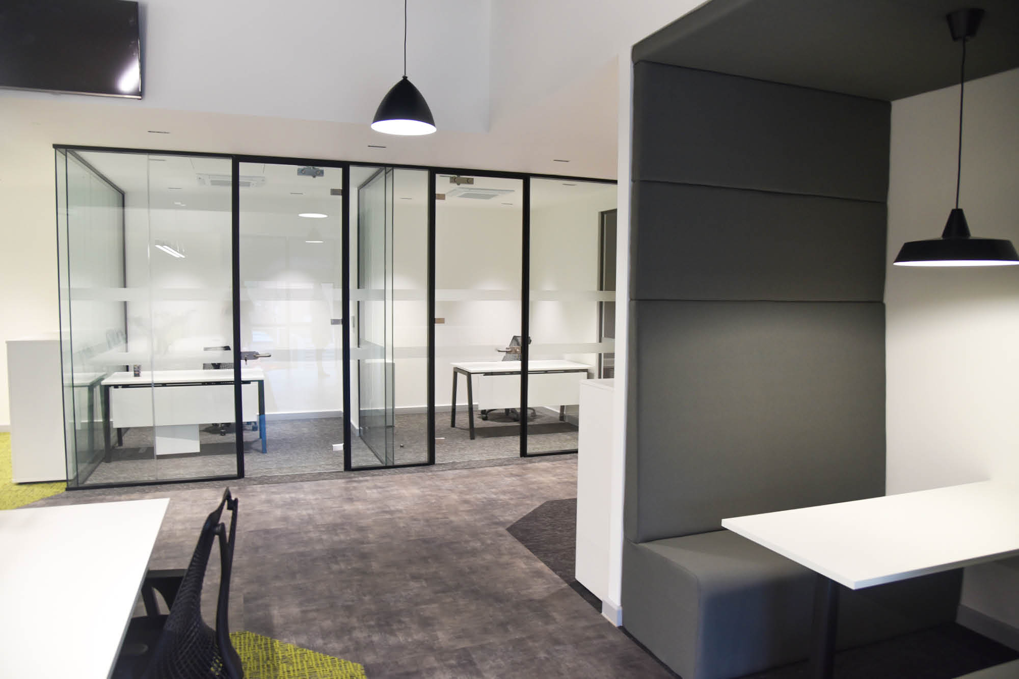 Modern office space with glass-walled meeting rooms. There are white tables and black chairs, along with a booth seating area under a hanging black lamp.