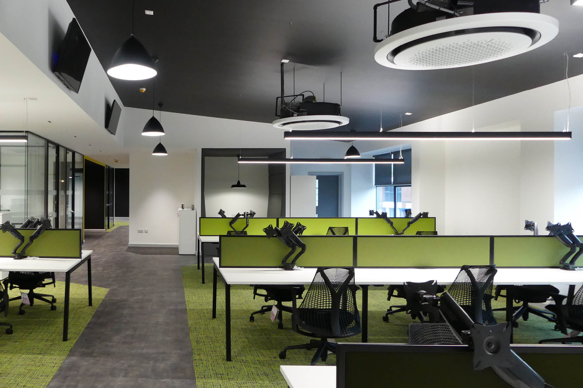 A modern office space with green dividers and black chairs at individual workstations. The ceiling features circular lighting fixtures.