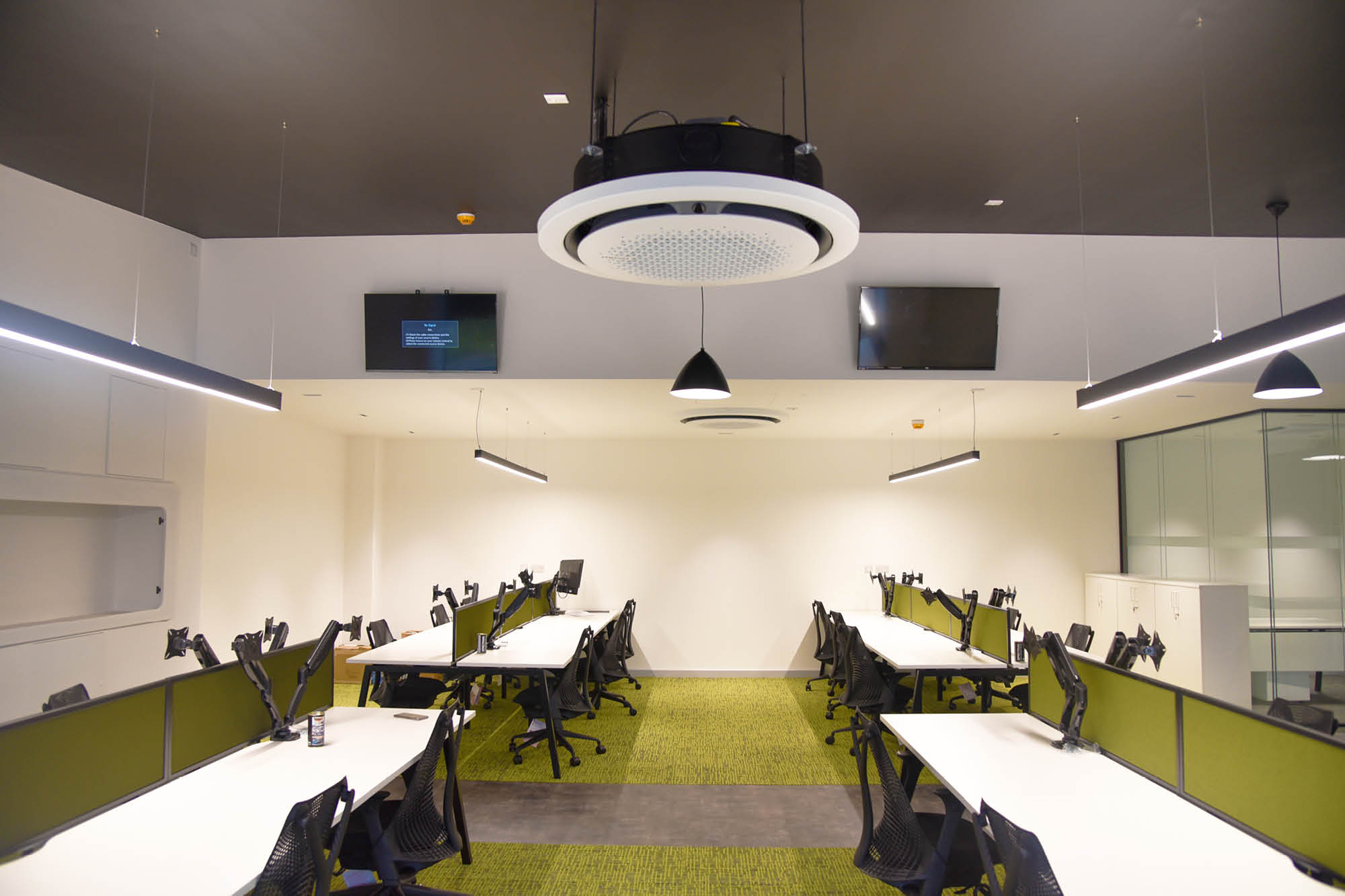 Modern office space with long tables and ergonomic chairs arranged in rows. Green carpeted floor and white walls with mounted TVs.