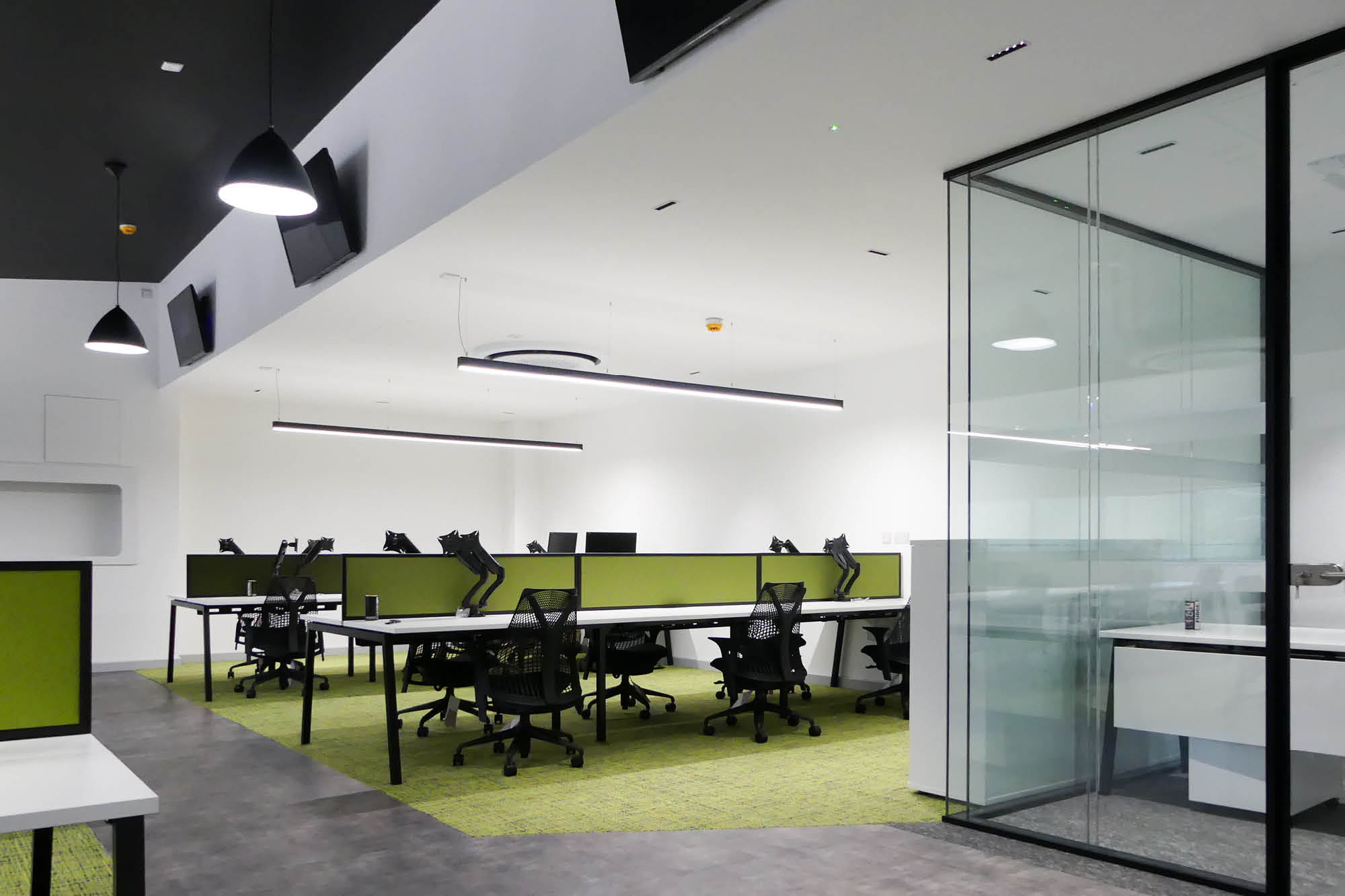 A modern office space with green-accented desks and black chairs arranged in rows. The room features sleek lighting.