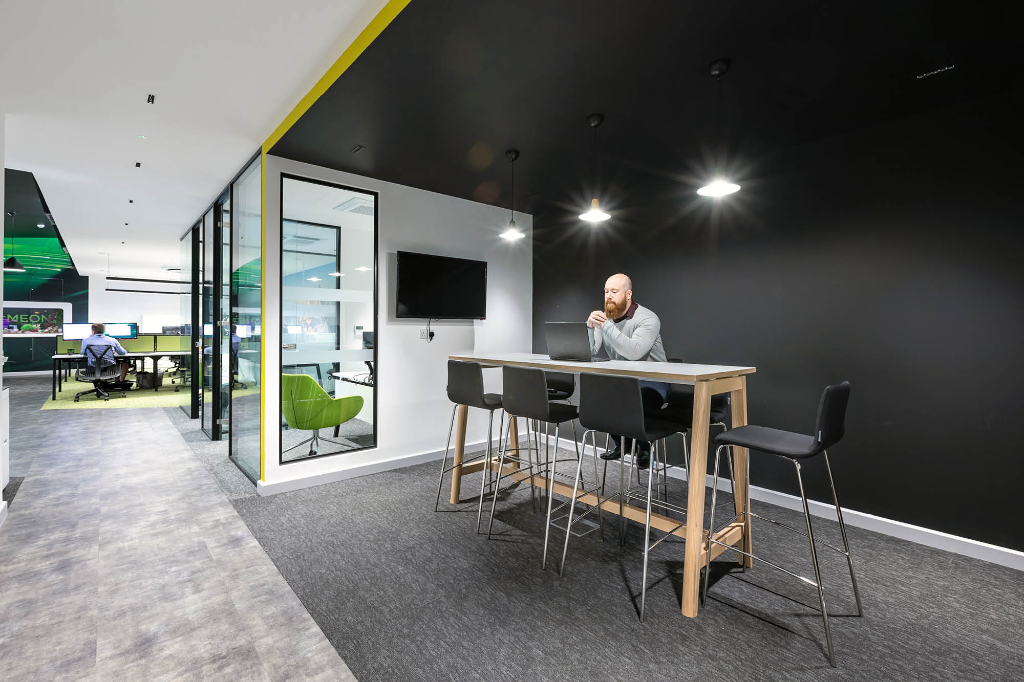A man sits at a high table in a modern office space with a black accent wall. The area has bar stools and overhead lights.