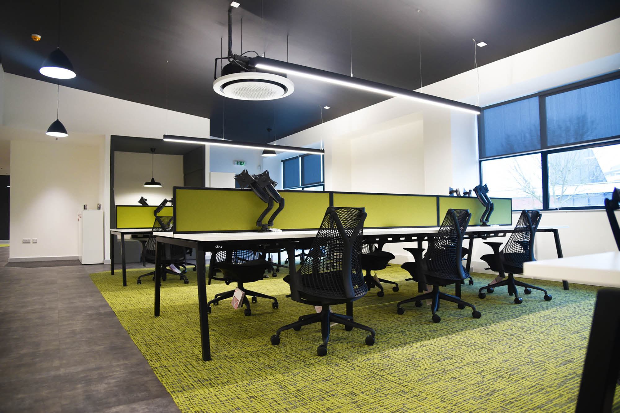 A modern office space with green carpet and black office chairs around a long table. Each station has a monitor mounted on an arm.