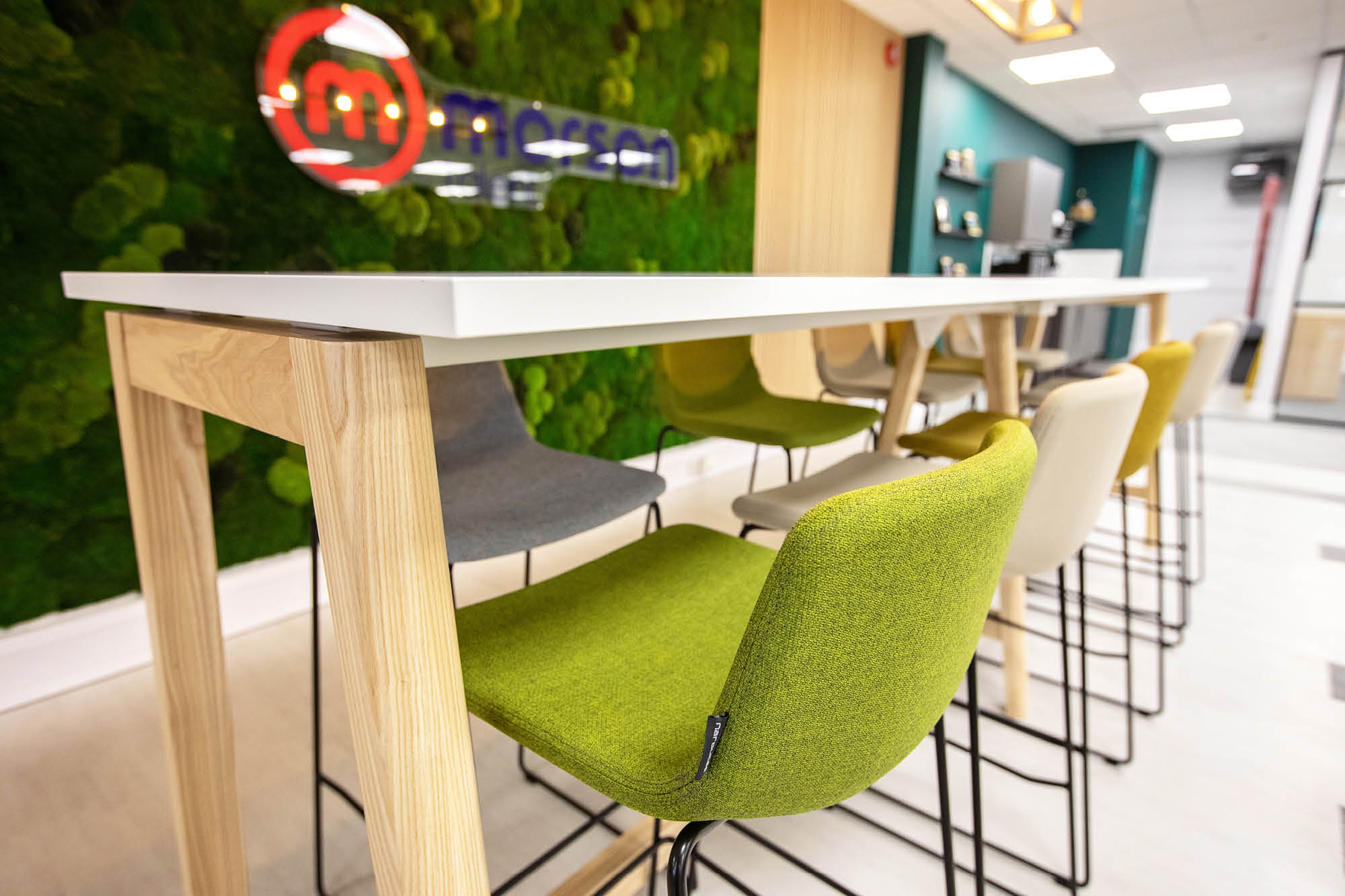 Modern office space with a long white table and green, gray, and beige chairs. The wall features a logo and green moss decoration.