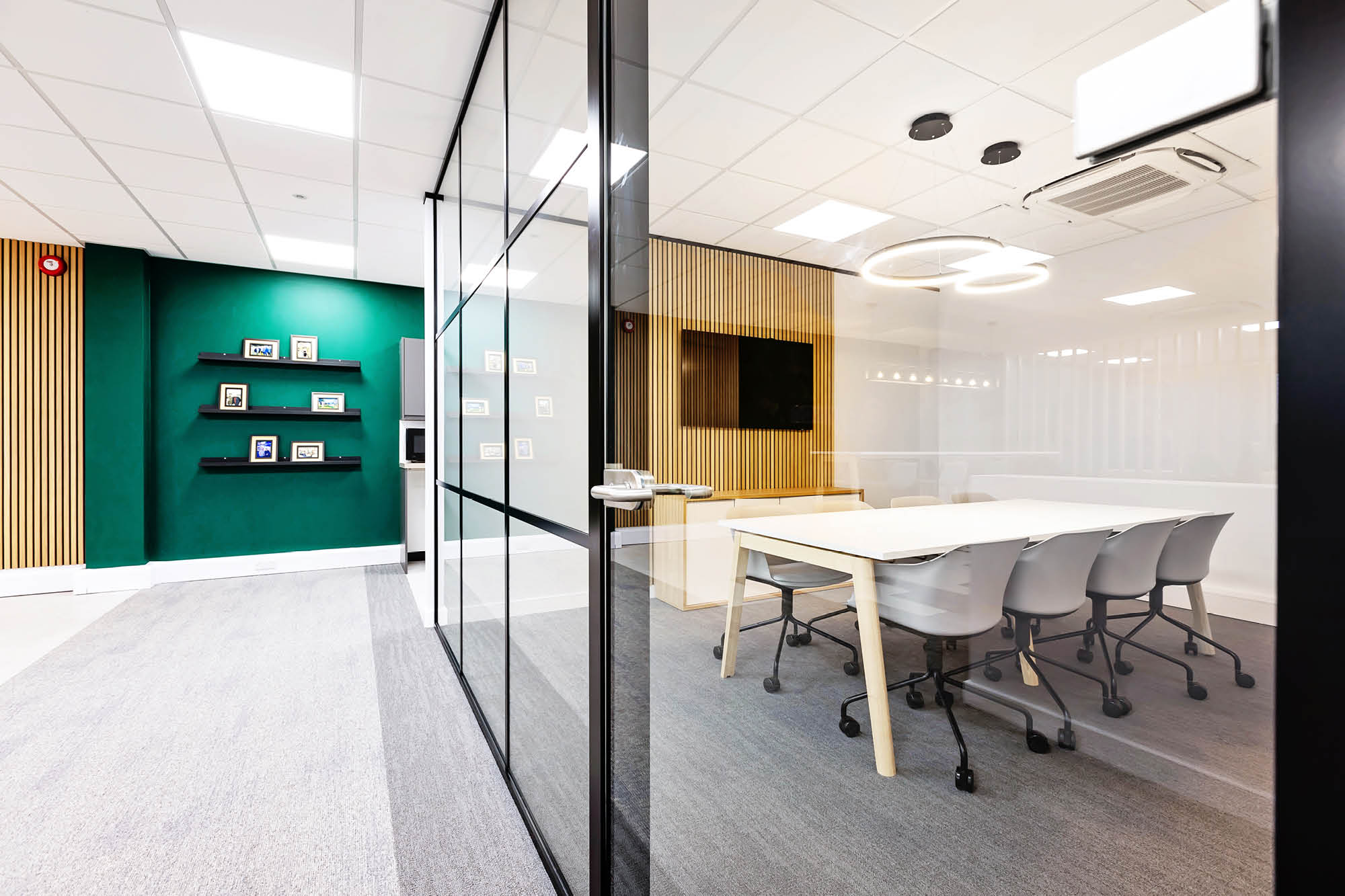 A modern conference room with a glass wall, featuring a large wooden table and six gray chairs. The room has a green accent wall.