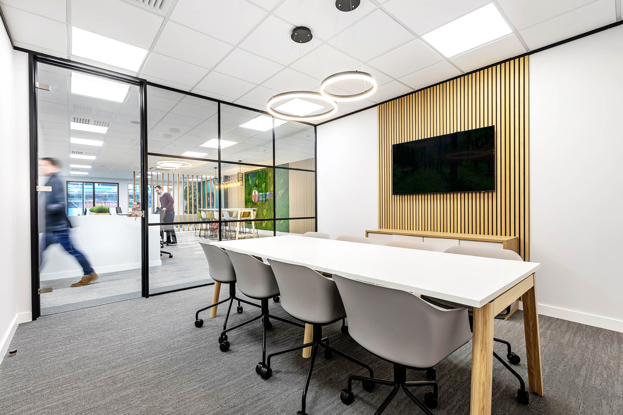 Modern office meeting room with a large white table, several grey chairs, and a wall-mounted TV.