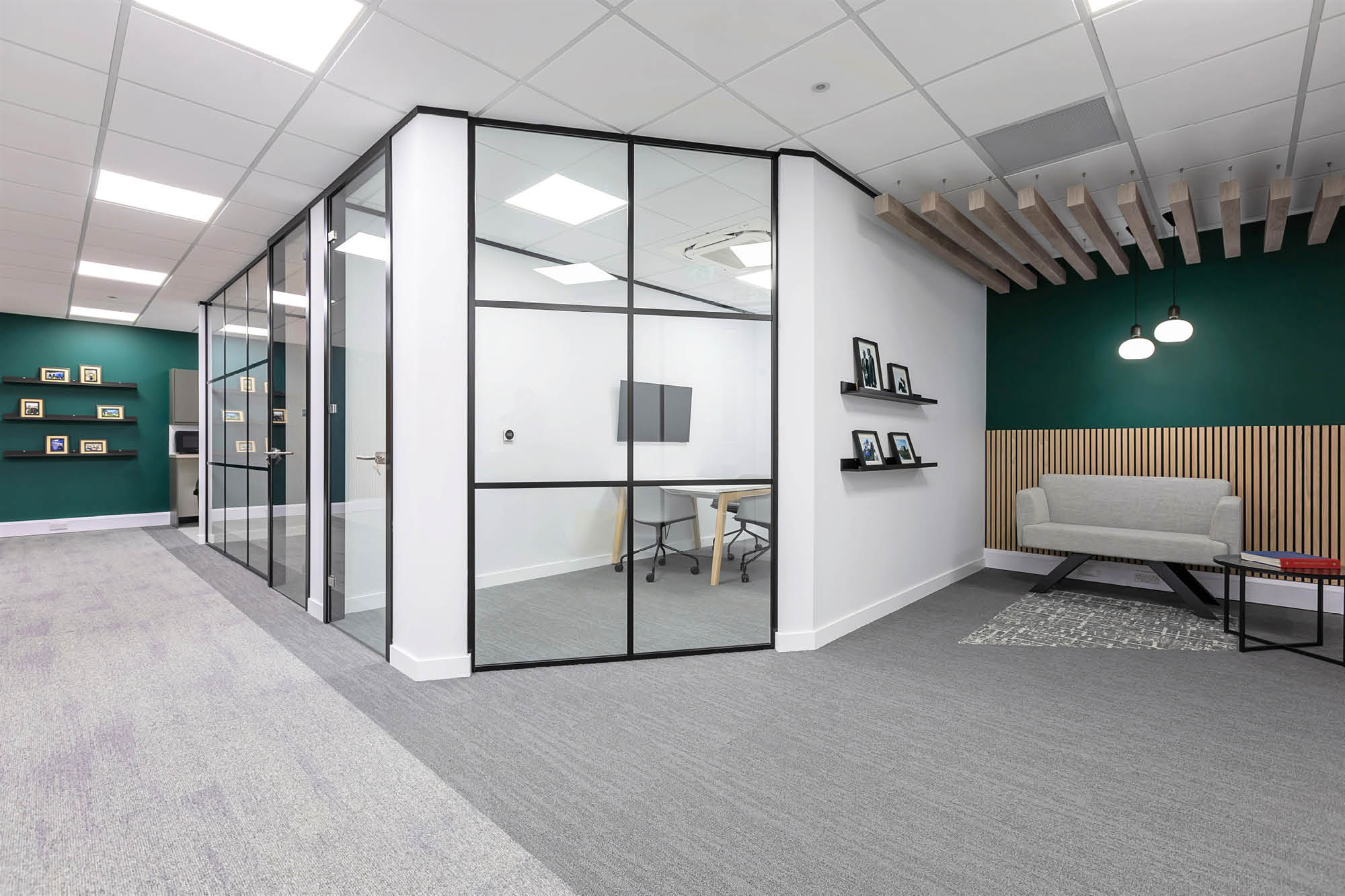 Modern office space with glass-walled meeting rooms, green and white walls, two shelves displaying decor, a grey carpet.