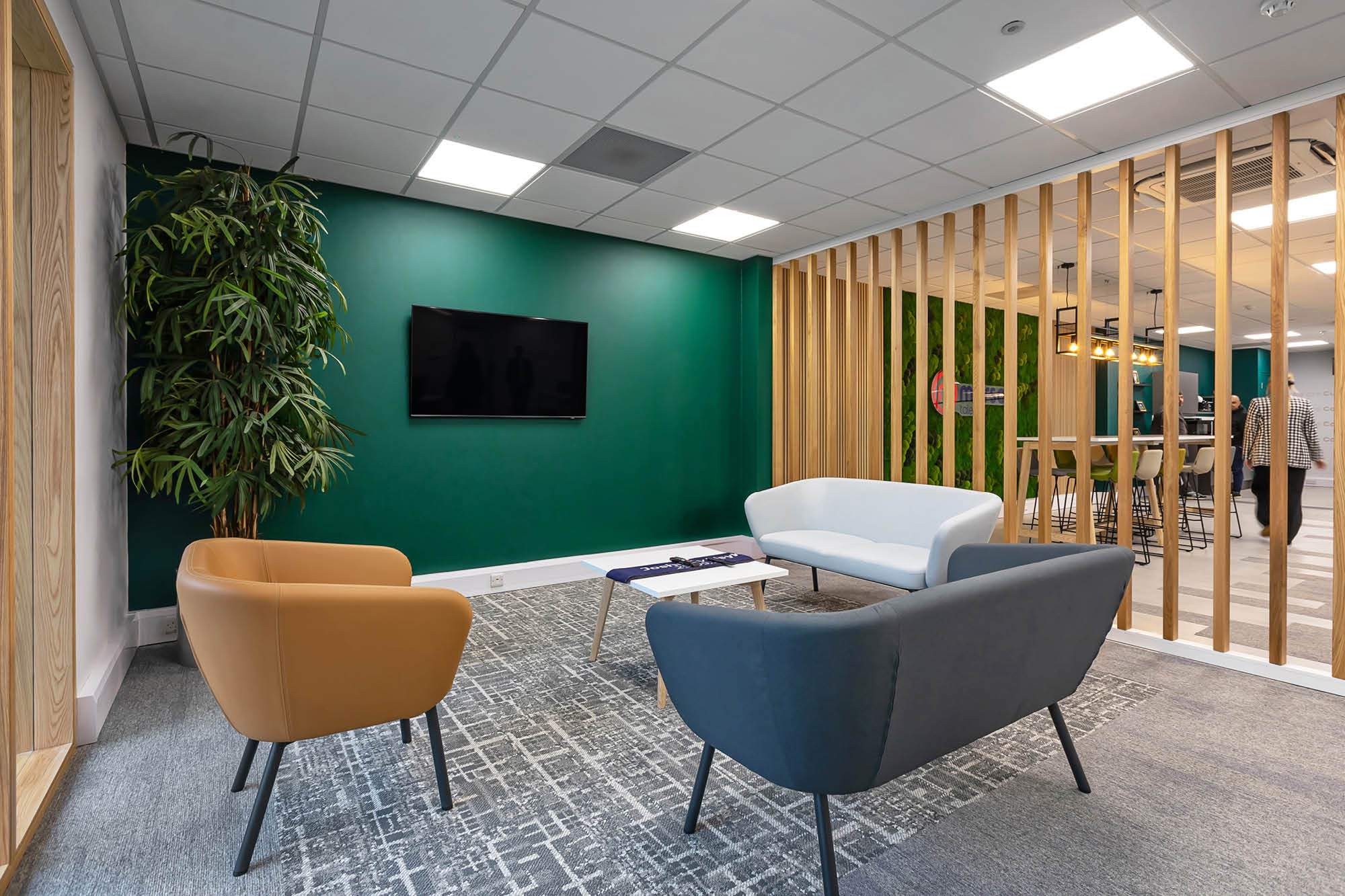 Modern office waiting area with green accent wall, mounted TV, and three chairs - one brown, one white, and one grey.
