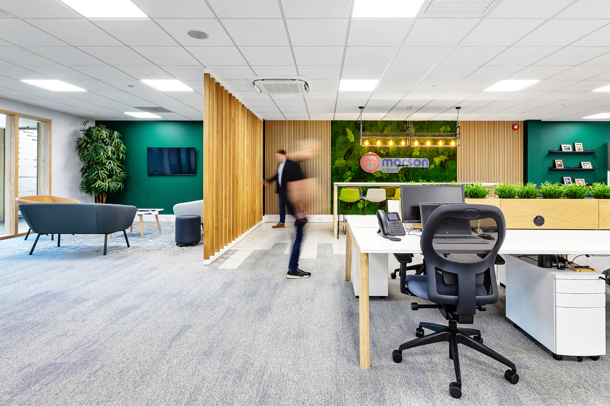 A modern office with a green wall and wooden slat dividers. Theres a blurred figure walking by. White desks with black chairs are in the foreground.