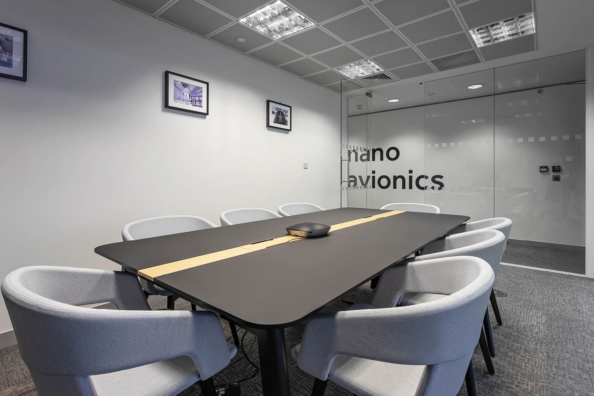 Modern conference room with a long black table surrounded by eight light gray chairs. The room has white walls, a glass door with partial text nano avionics.