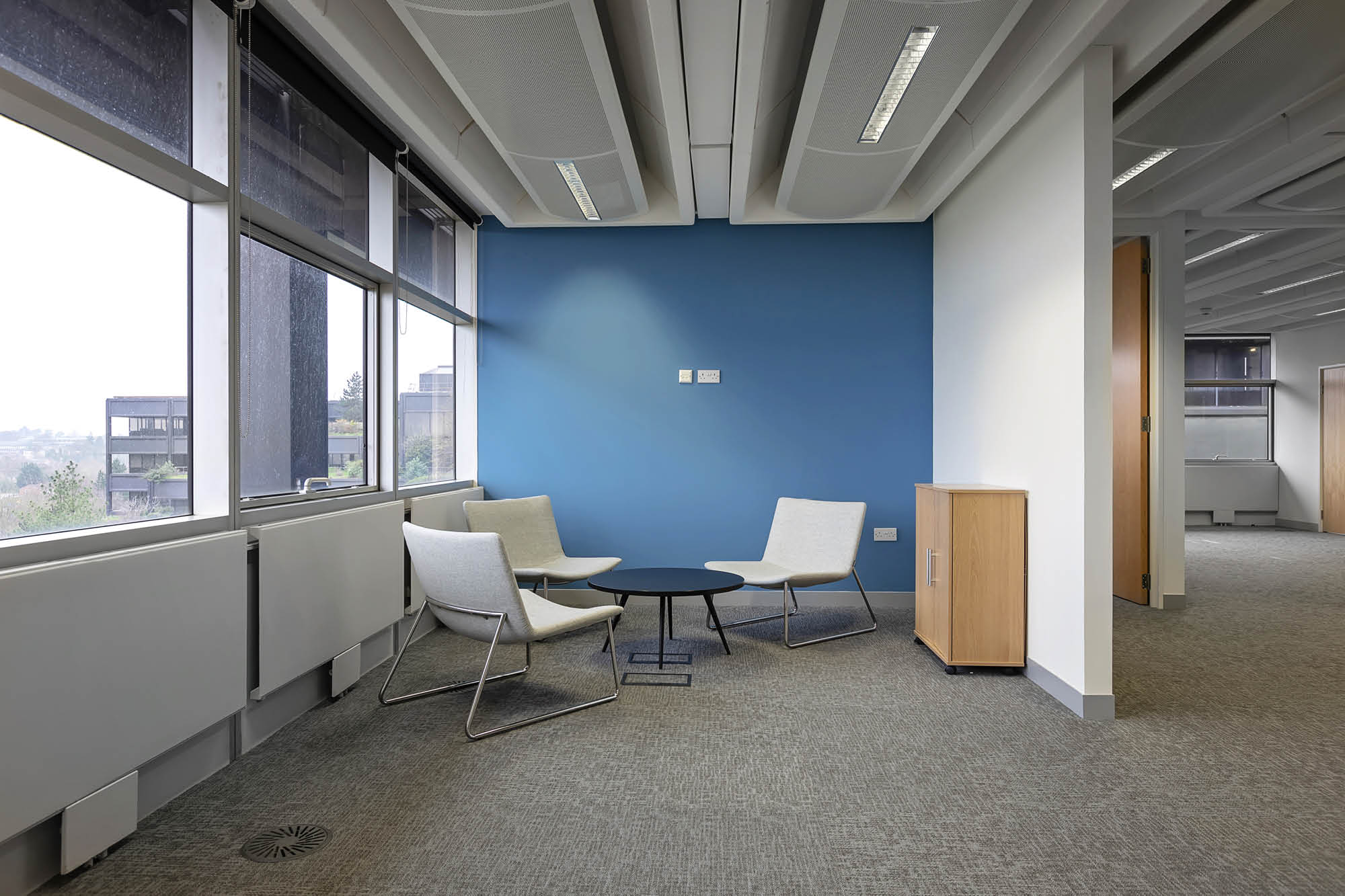 Modern office corner with three sleek chairs around a round black table on a gray carpet. The wall is painted blue.