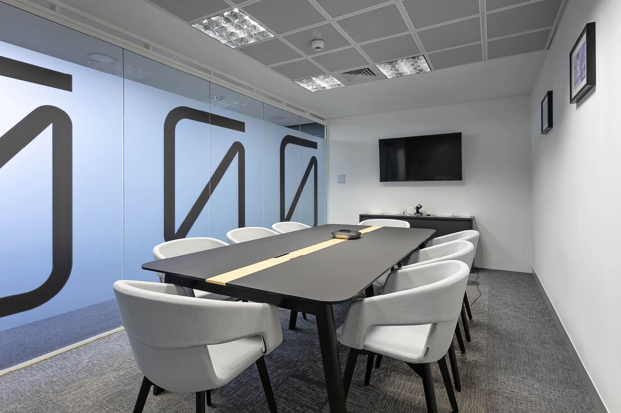 A modern conference room with a long black table surrounded by six white chairs. Theres a flat-screen TV mounted on the wall.